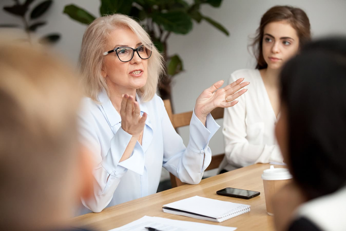 woman with elbows on table holding out hands while making a point in a meeting around a conference table
