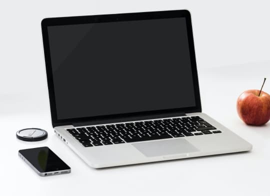 Silver laptop shown open surrounded by phone and apple