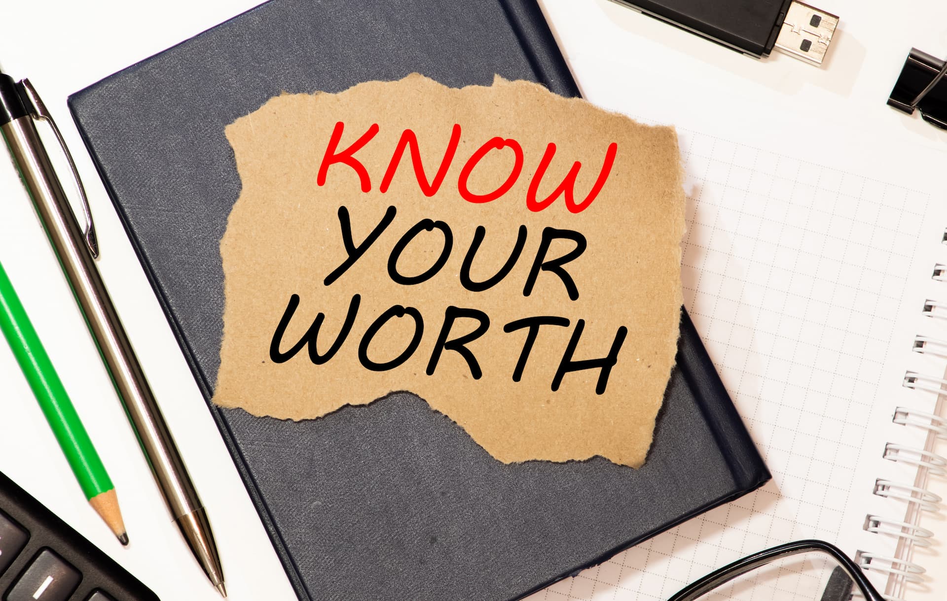 Know Your Worth written on torn brown paper sitting on top of a notebook and other office supplies on a desk.