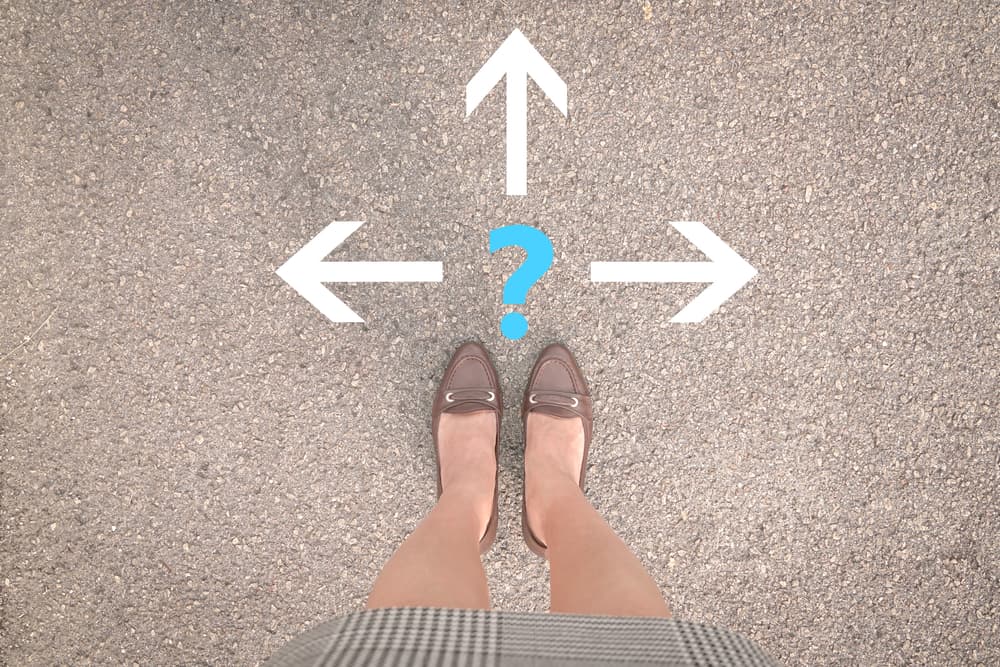 Businesswoman standing on street looking down at a question mark with chalk arrows drawn going left, right and forward, contemplating a direction decision.