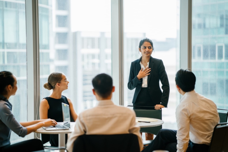 Office meeting where four team members pay close attention to their leader, who is touching her chest, signaling that she’s opening up about her concerns.