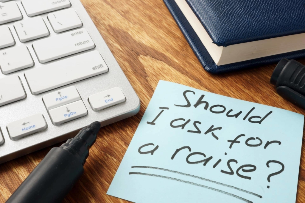 How to ask for a raise: Office desk with office items scattered and a post it note that reads, Should I ask for a raise?