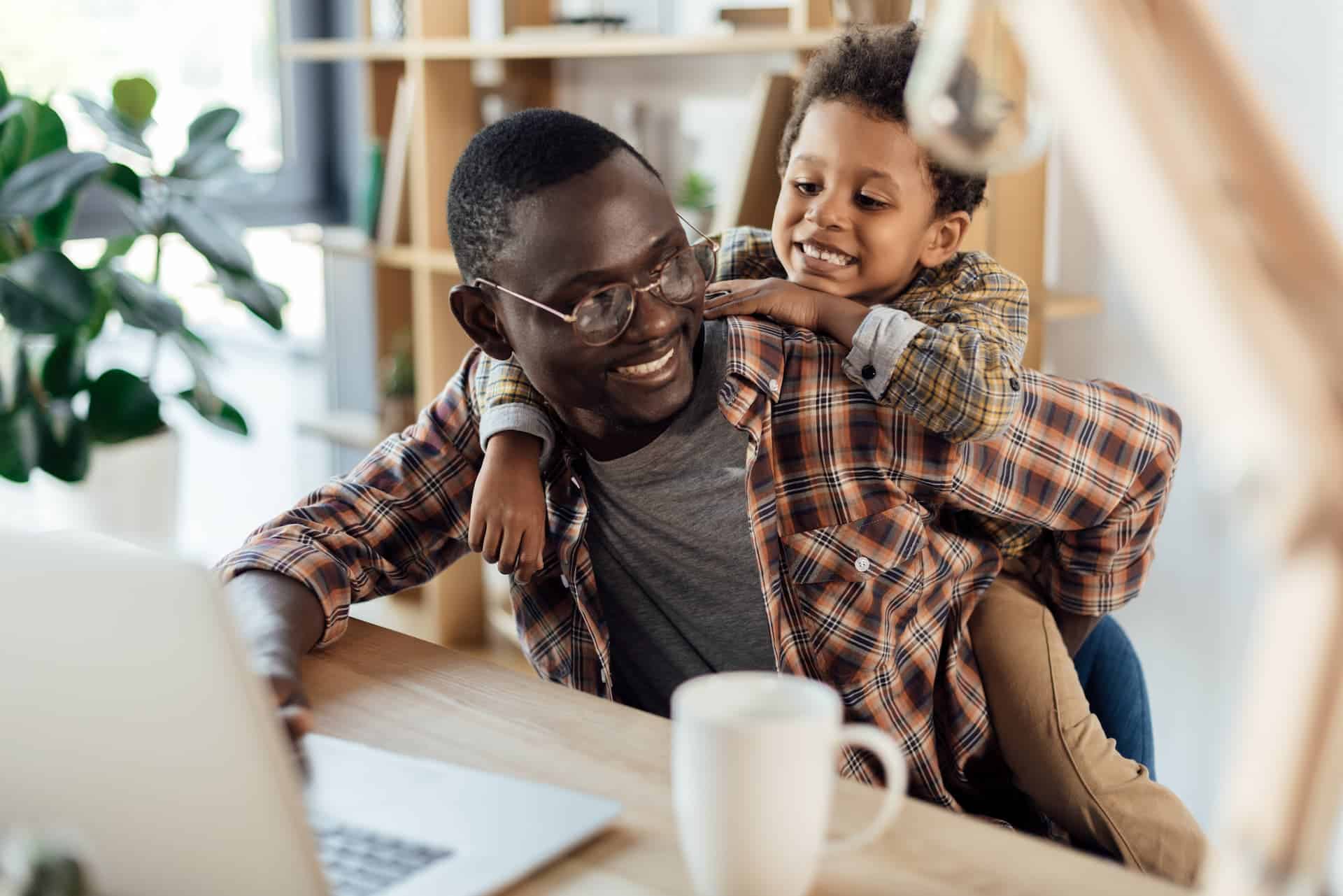 A father on his computer hugging his young son who jump on his back.