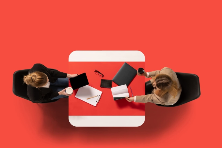 Two young women sit at common table with documents and conduct interview against red studio background.