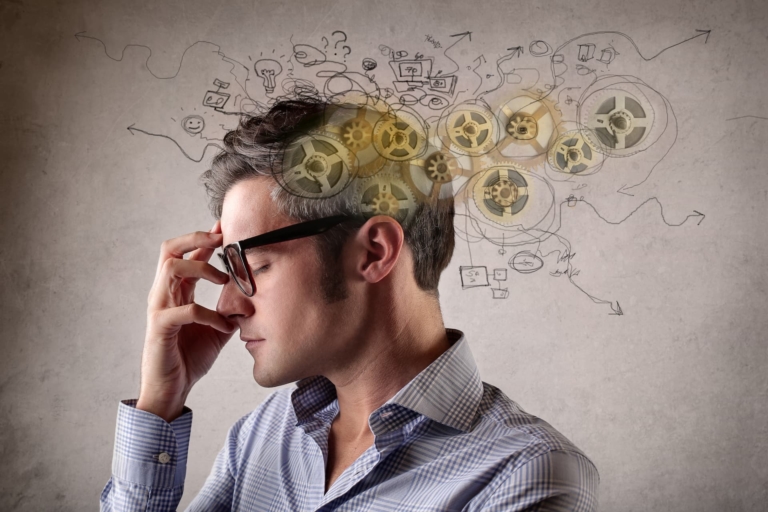 A man with closed eyes, hand to his forehead, with cogs and scattered line drawings of arrows, circles, and doodles representing his thoughts, surrounding the top of his head.