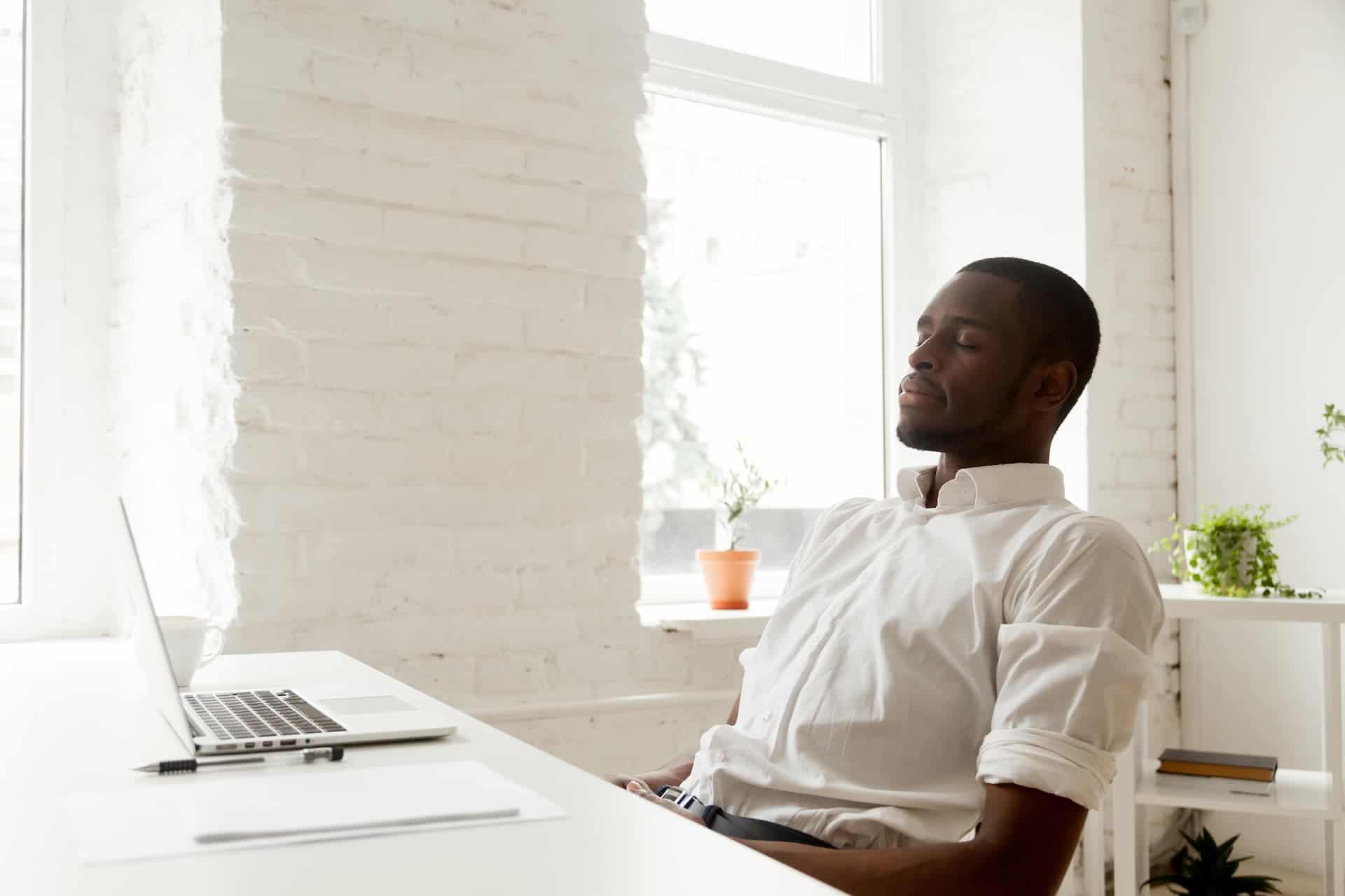 A man seated in a chair at work with his eyes closed, practicing mindfulness by focusing on his breath.