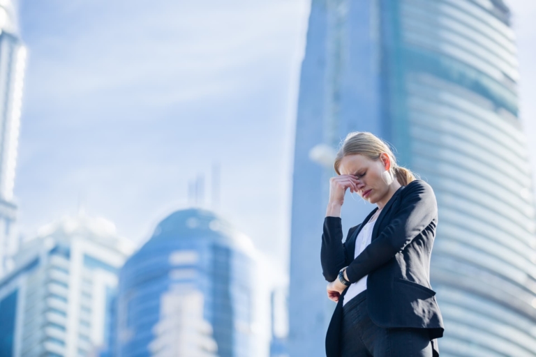 A woman in work attire standing before a backdrop of modern city buildings, her hand on her face in a facepalm gesture, conveying a sense of regret or frustration in a professional context.