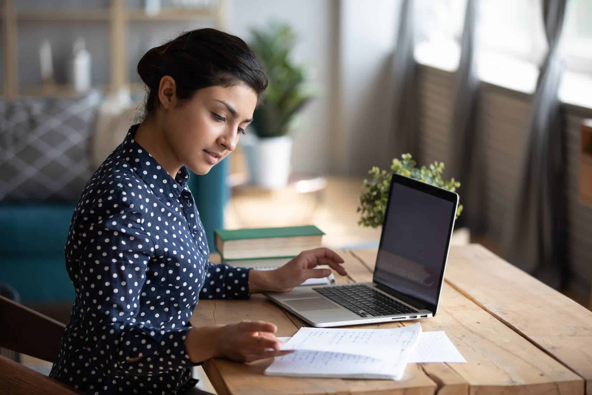 A woman happily engrossed in her work, typing on a laptop while referencing a notebook, representing the state of flow and deep engagement.
