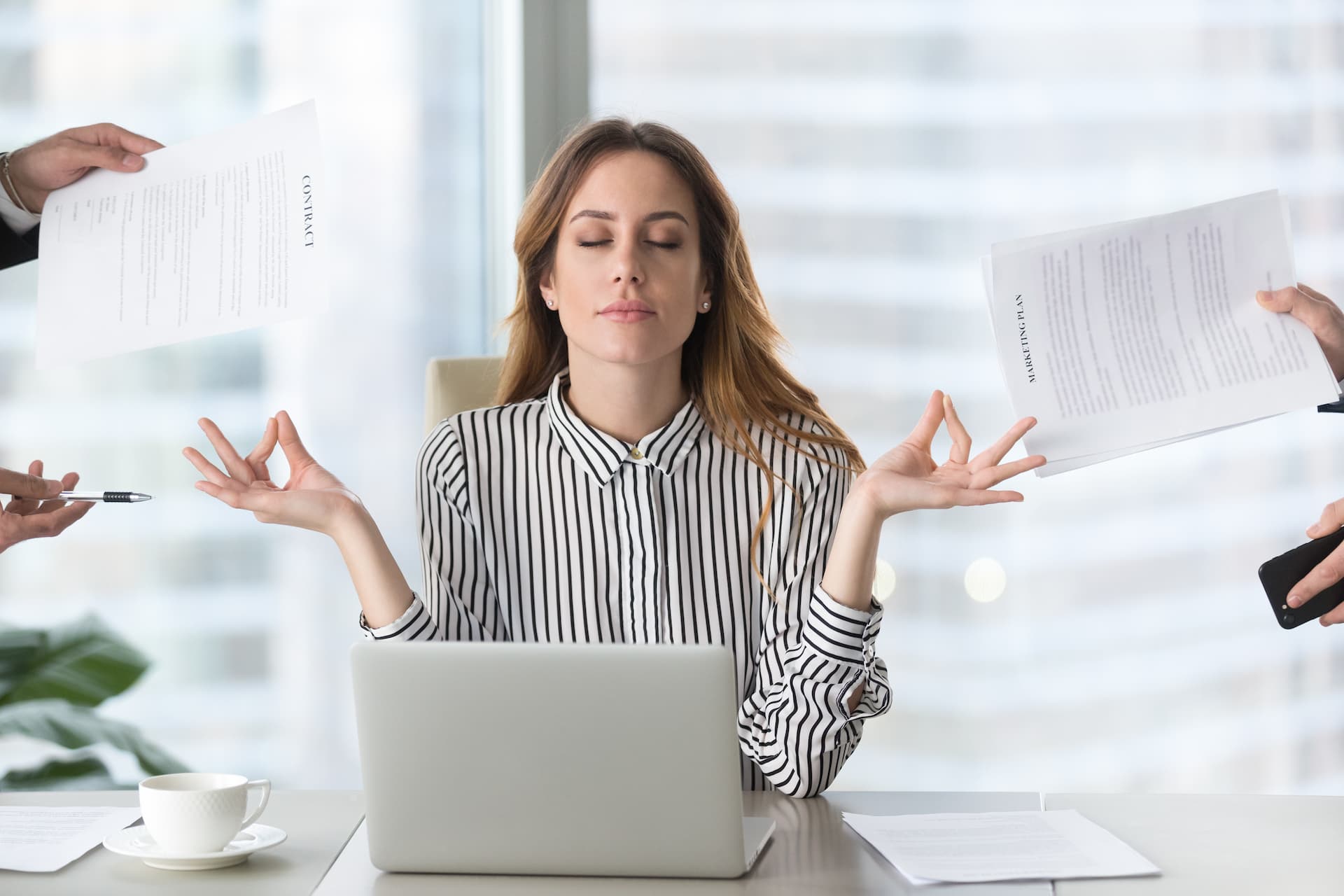 Calm female executive meditating in front of her laptop while coworkers push paperwork and assignments toward her.