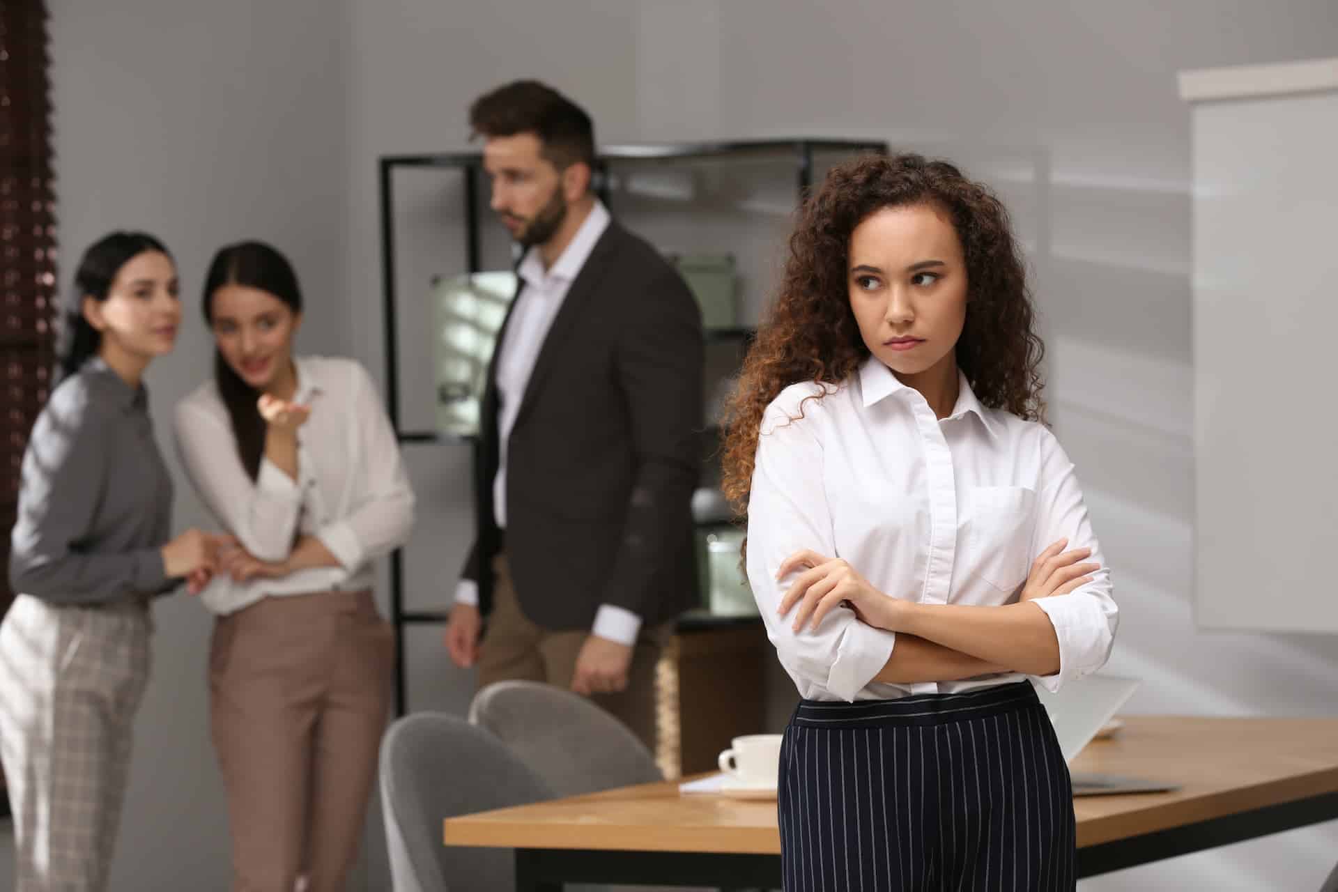 A woman stands alone outside a group, appearing sad, while others engage in conversation, hinting at the isolating effects of office politics and gossip in the workplace.
