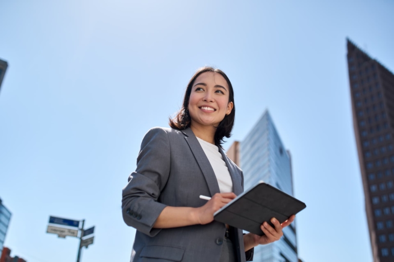 Smiling woman in a cityscape, confidently holding an iPad and Apple Pencil, exuding professionalism and entrepreneurial spirit.