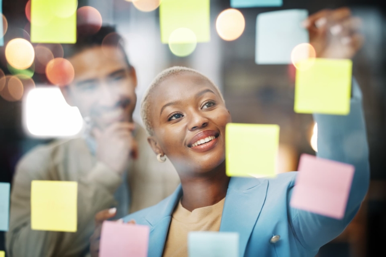 A professional woman in an office pasting Post-it notes on a see-through board illustrating the creative process of brainstorming and idea generation.
