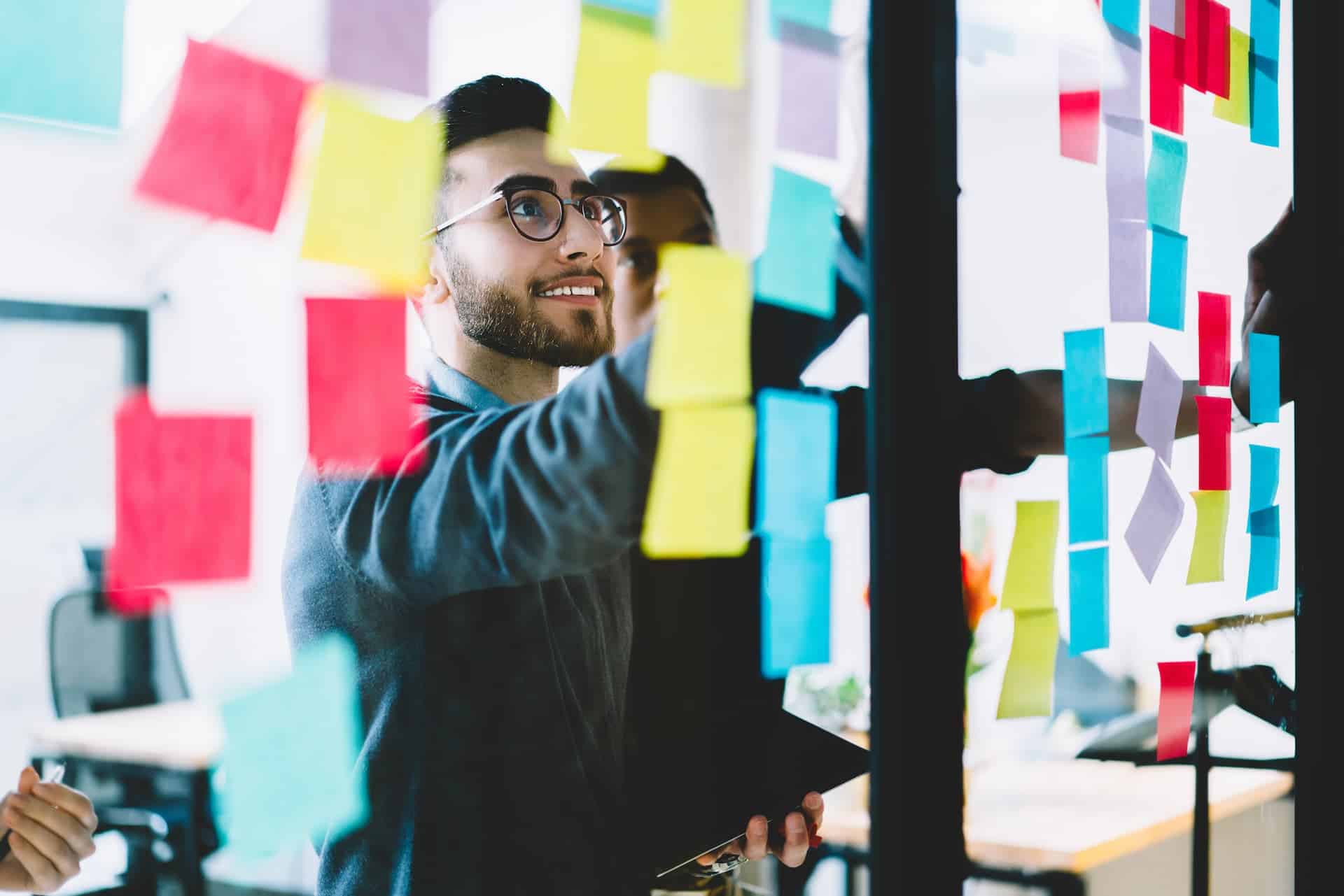 Young men collaborating in an office setting, brainstorming and organizing ideas with colorful post-it notes on a glass door, embodying the creative and collaborative spirit of entrepreneurship.
