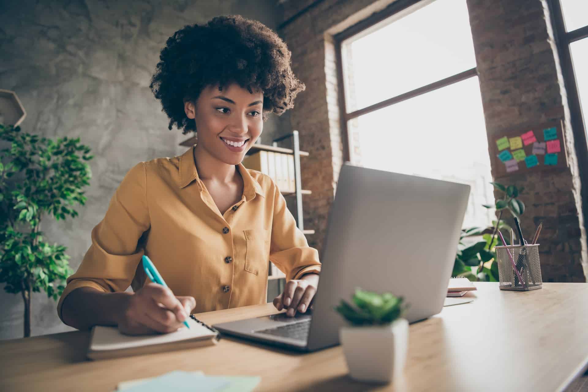 Woman sitting at a desk, using her laptop to research job opportunities while taking notes in a notebook.