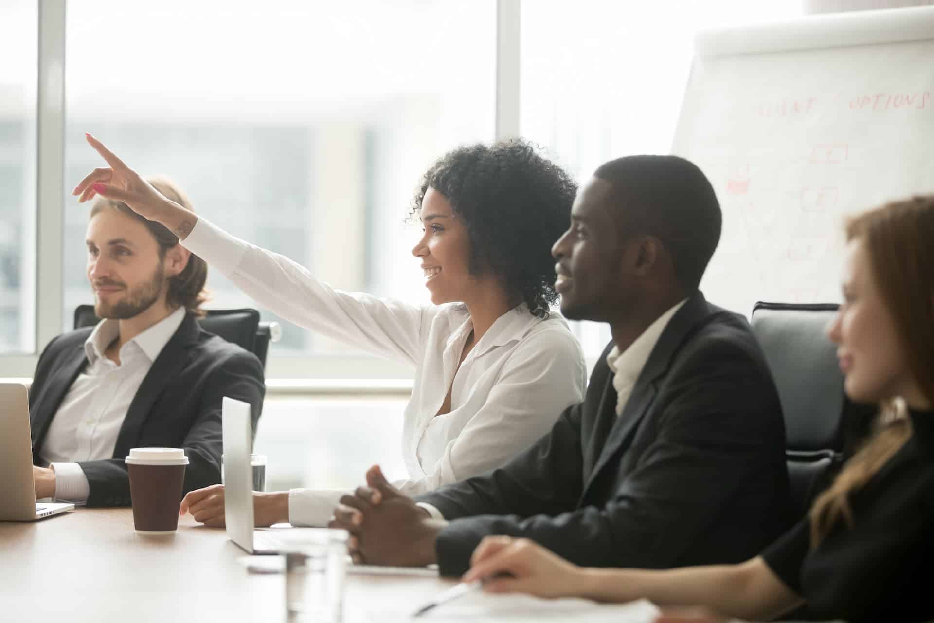 Employees engaged in a small meeting, with one raising her hand, symbolizing an open culture of curiosity and active participation.