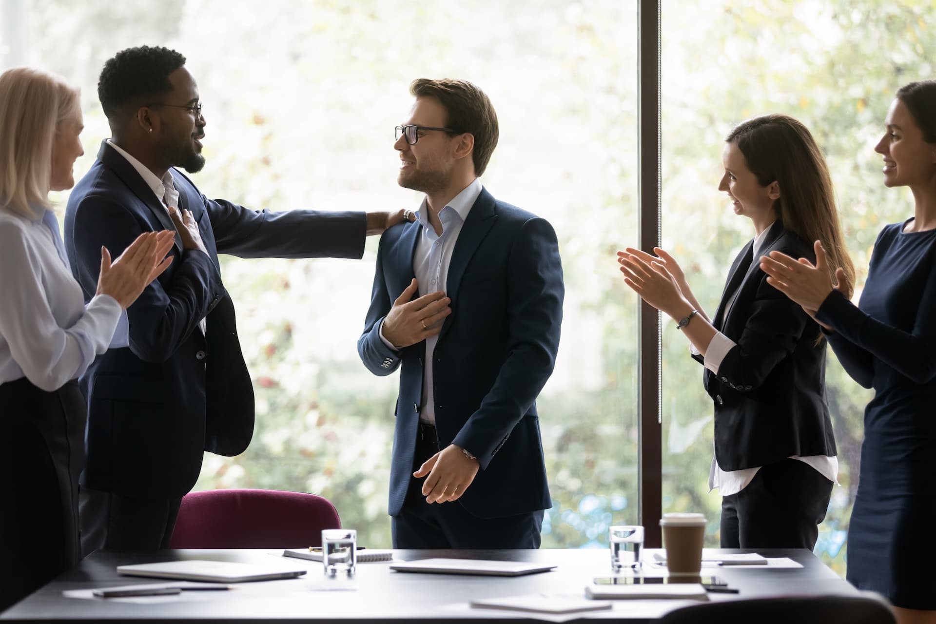 Business men in women in conference room congratulating colleague by clapping and touching shoulder.