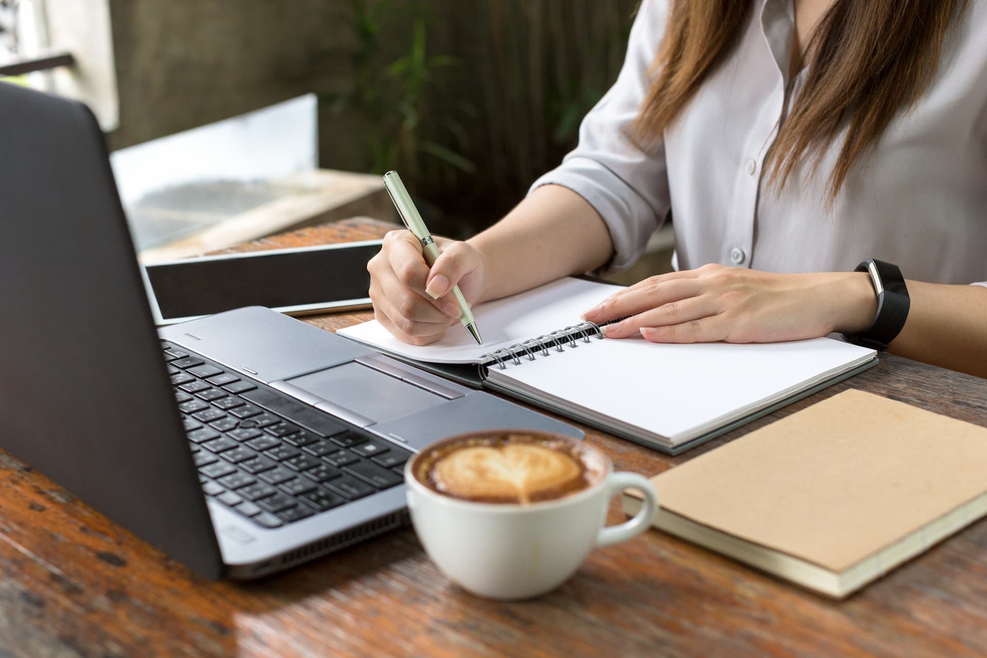 Close-up of a woman's hand taking notes in a notebook while referencing a computer screen, symbolizing personalized coaching services.