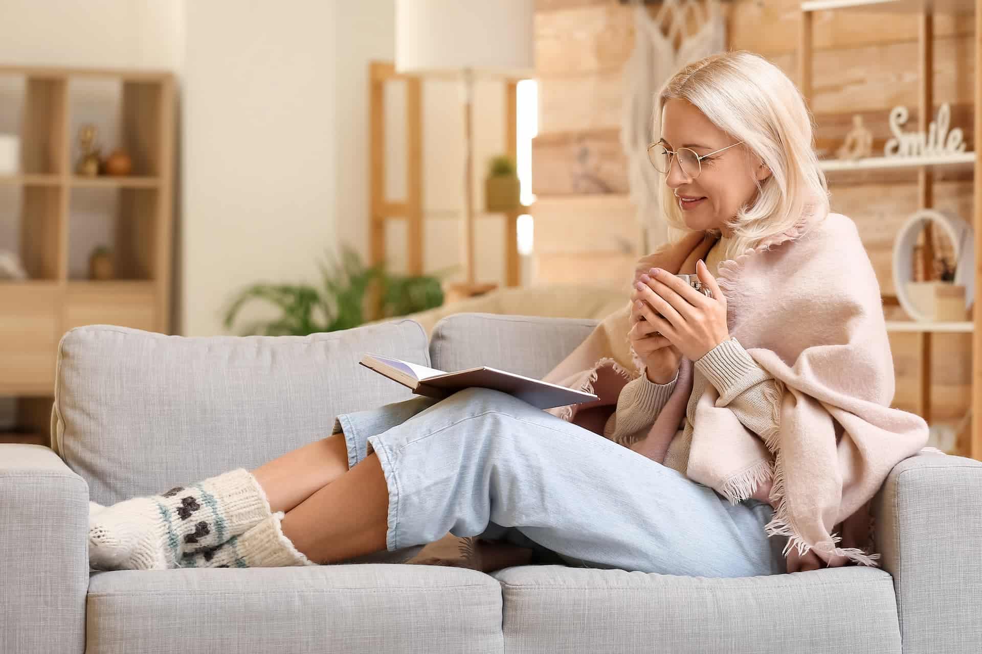 Woman sitting on a cozy coach, reading a book and drinking tea, disconnecting from work and prioritizing her self-care. 