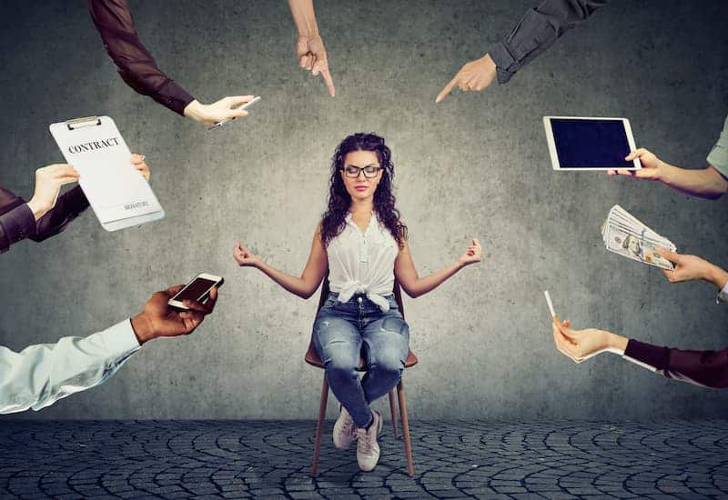 A woman practicing mindfulness exercises on a chair, surrounded by hands holding papers, laptops, and other devices, showcasing the need to set boundaries at work.