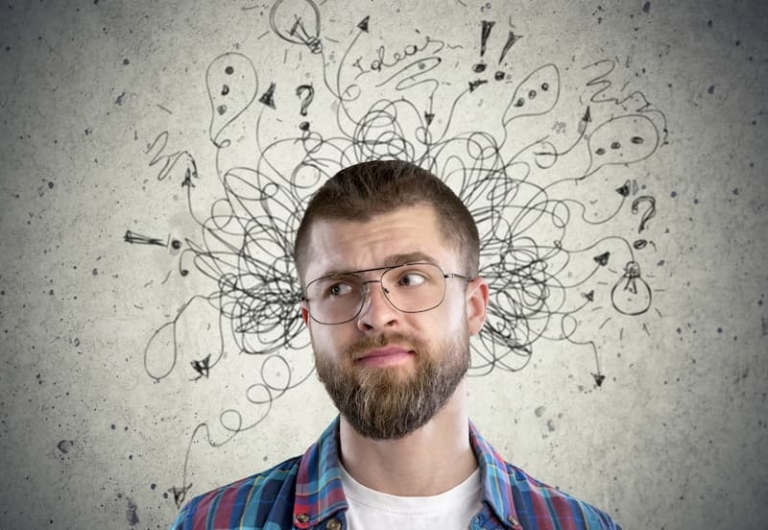 Closeup of a man wearing glasses, deep in thought, surrounded by a halo of scribbles and drawings on the wall behind him, symbolizing intense critical thinking and creativity.
