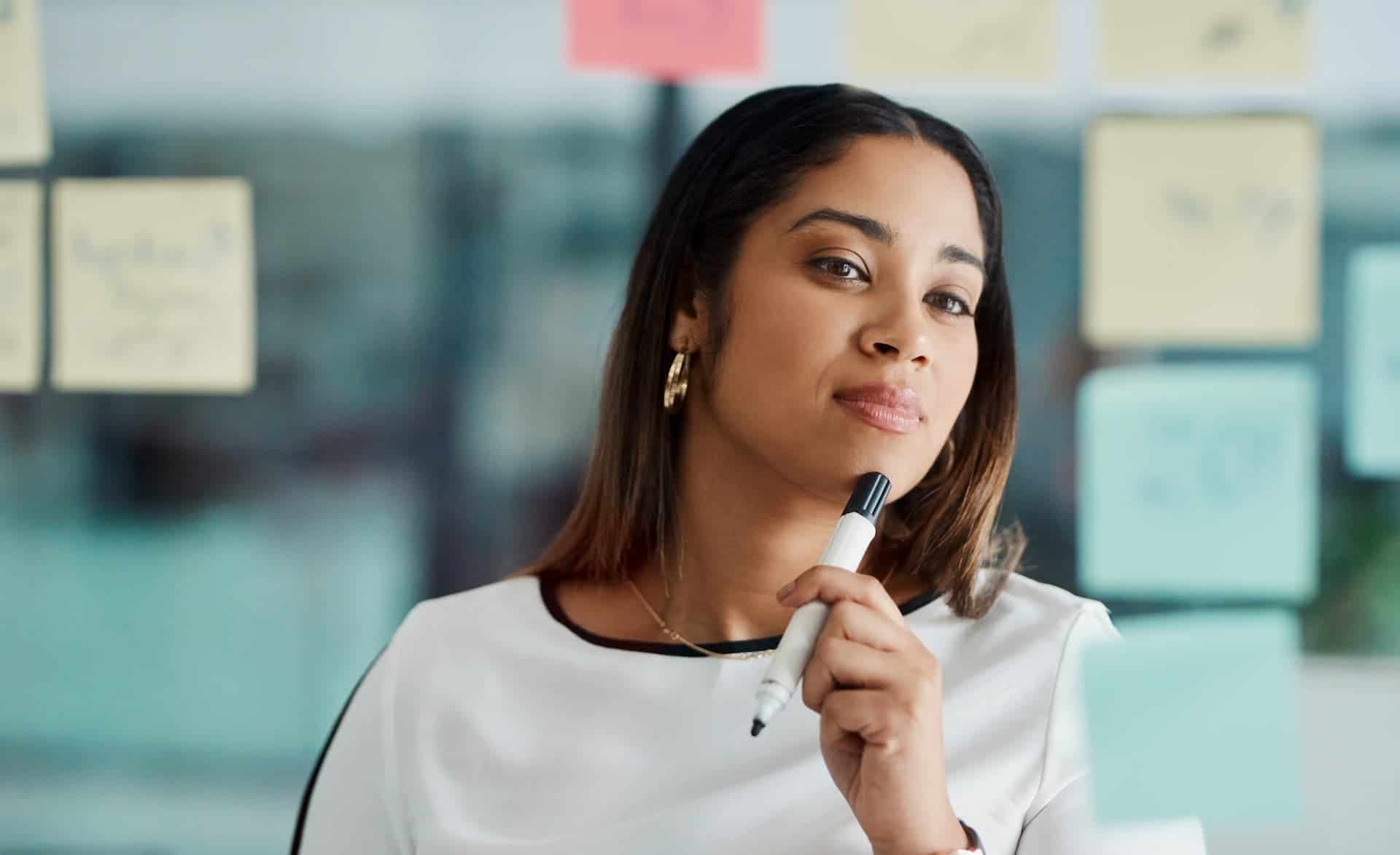 Woman in her office, staring at post-it notes, and holding a marker to her chin, thinking critically about her work projects.