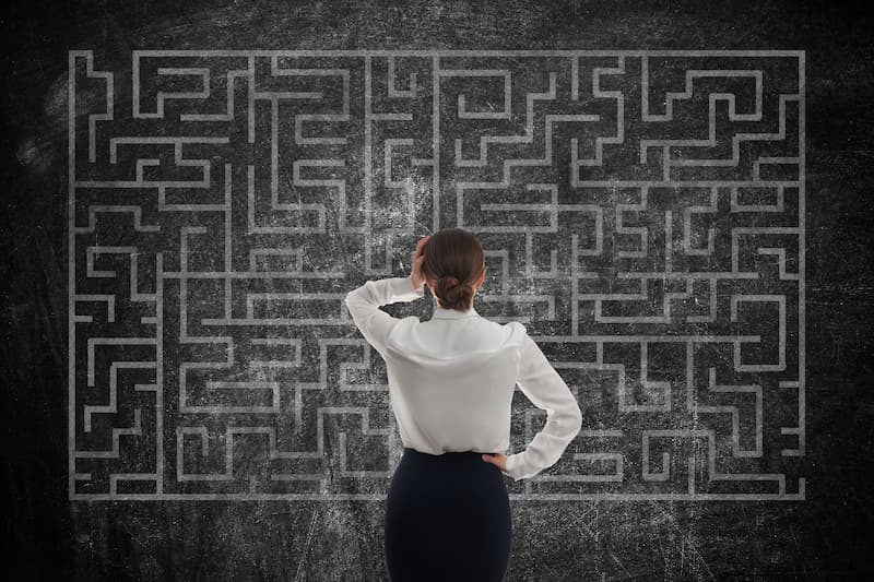 A woman in work attire standing in front of a chalkboard with a labyrinth drawing, appearing thoughtful and puzzled as she contemplates a problem-solving strategy.