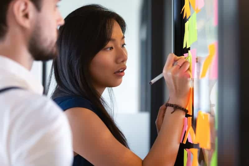 Two coworkers actively collaborating on a problem-solving task, using post-it notes to jot down ideas and affixing them to a glass door or window, demonstrating teamwork and brainstorming.