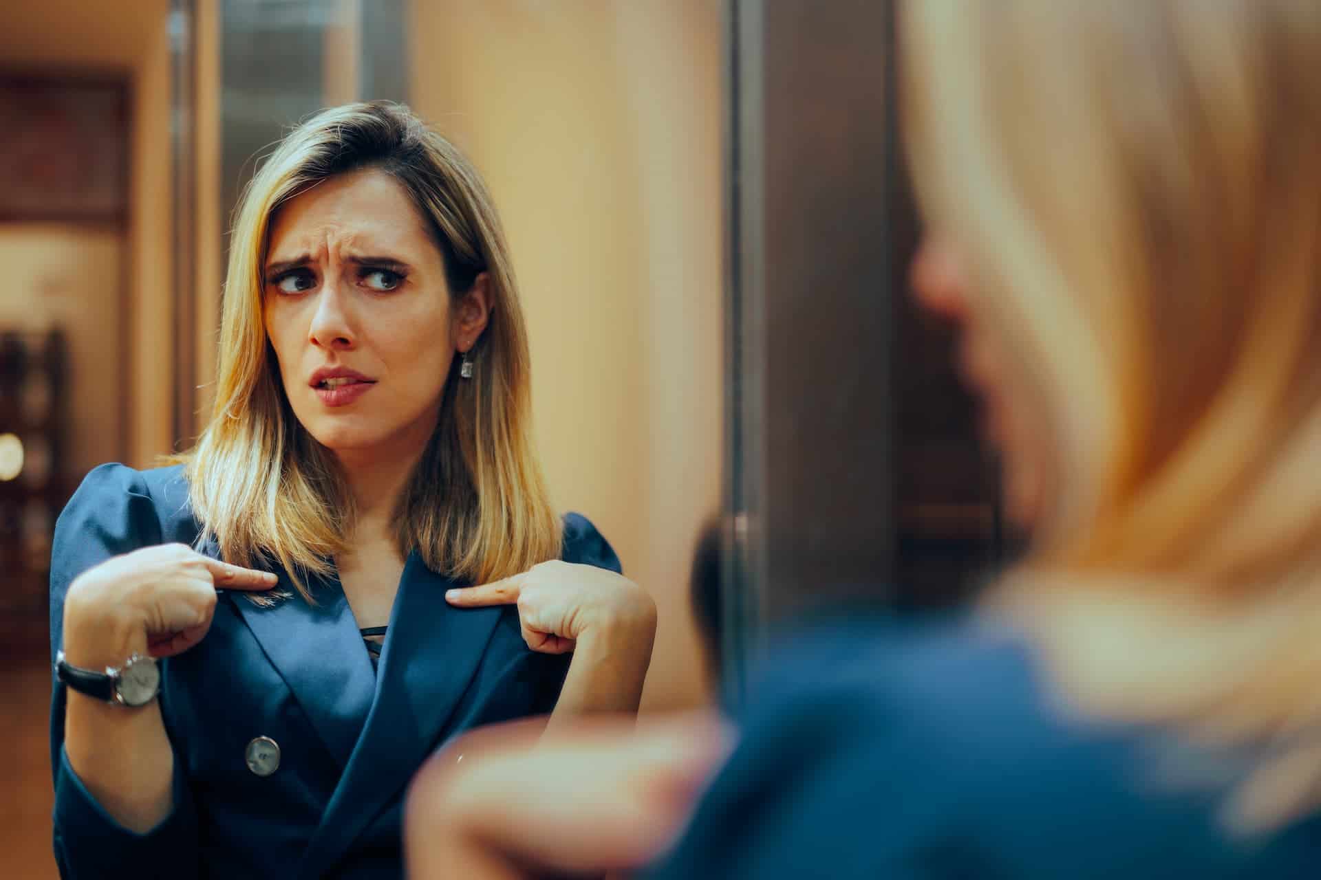 A woman in a suit looking at herself in the mirror, appearing insecure but working on building her self-confidence.