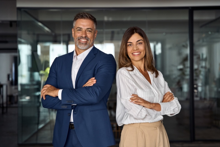 A man and woman in suits standing back-to-back in an office with their arms crossed, exuding confidence and conviction.