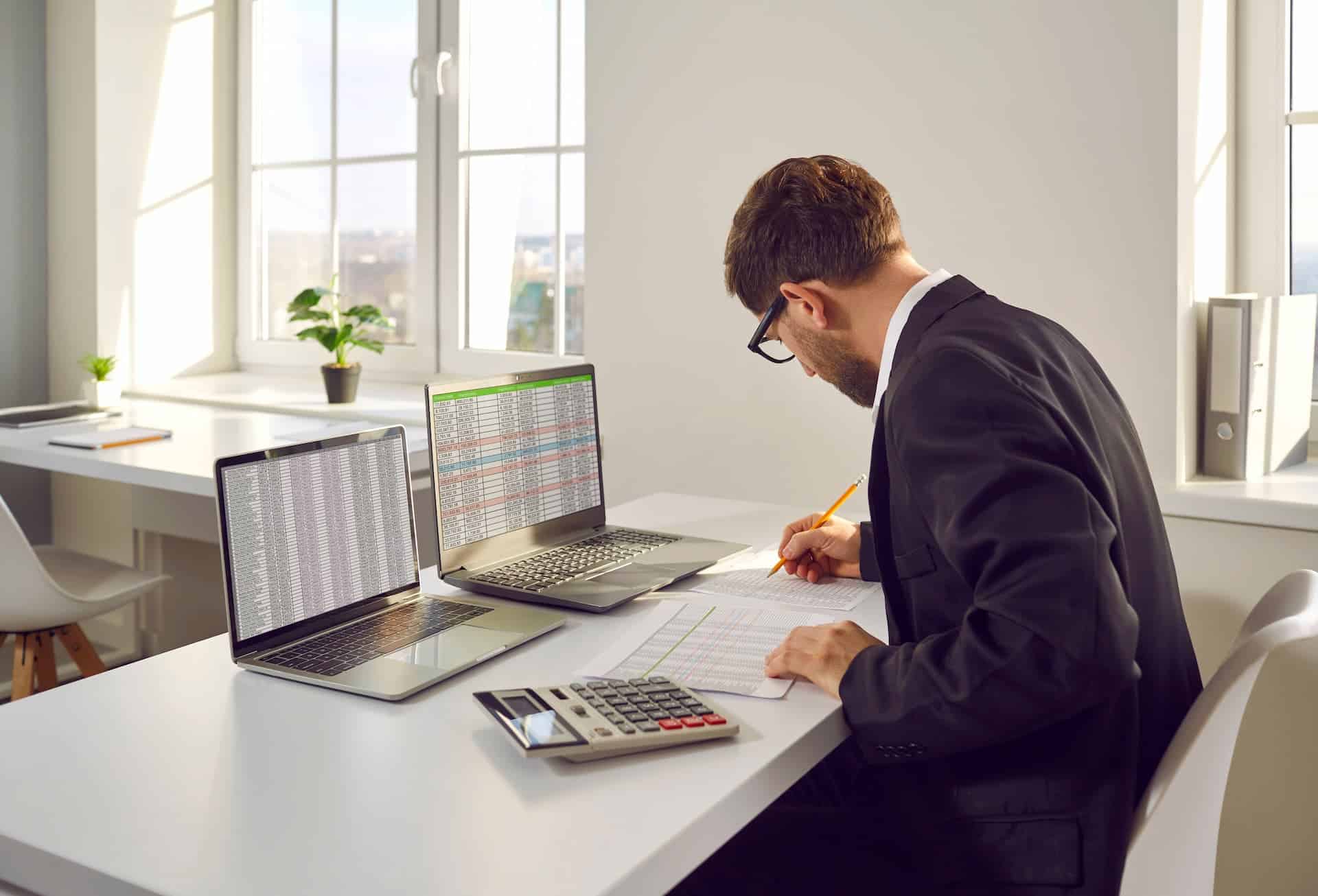 A man in an office with two laptops, a calculator, a pencil, and papers, diligently working with conviction.