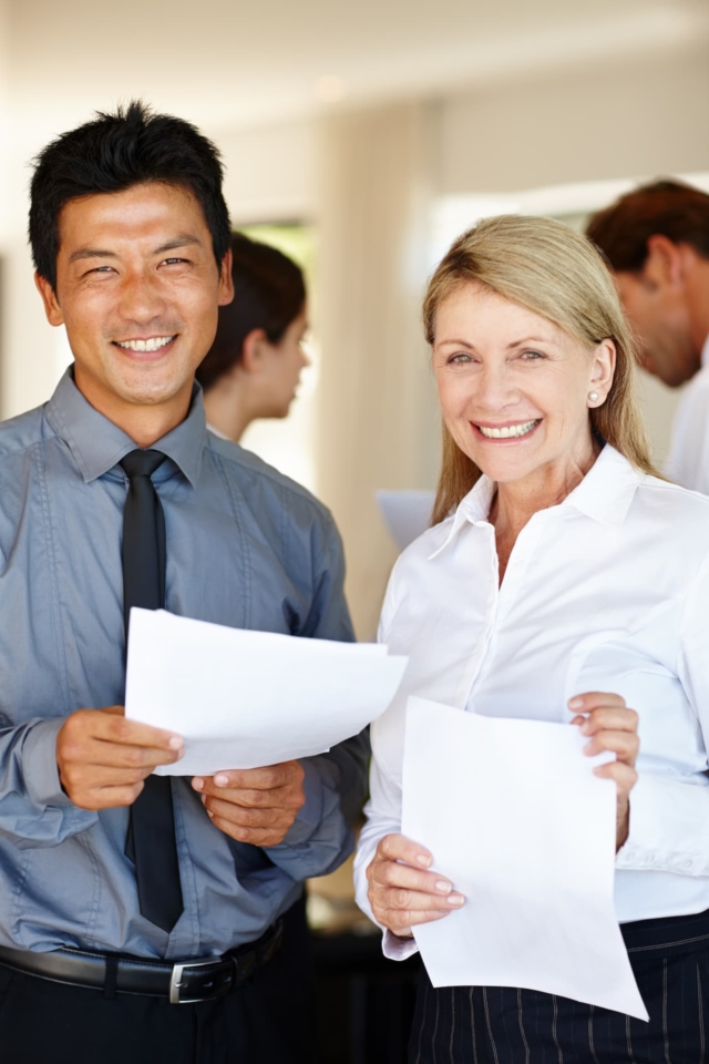 A mature woman and an Asian man stand side by side, smiling and holding papers, representing a diverse and collaborative team, with two other people engaged in conversation in the background.