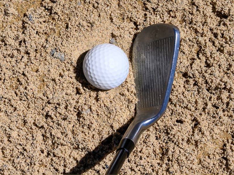 Golf ball in a sand bunker with a sand wedge poised for a shot.