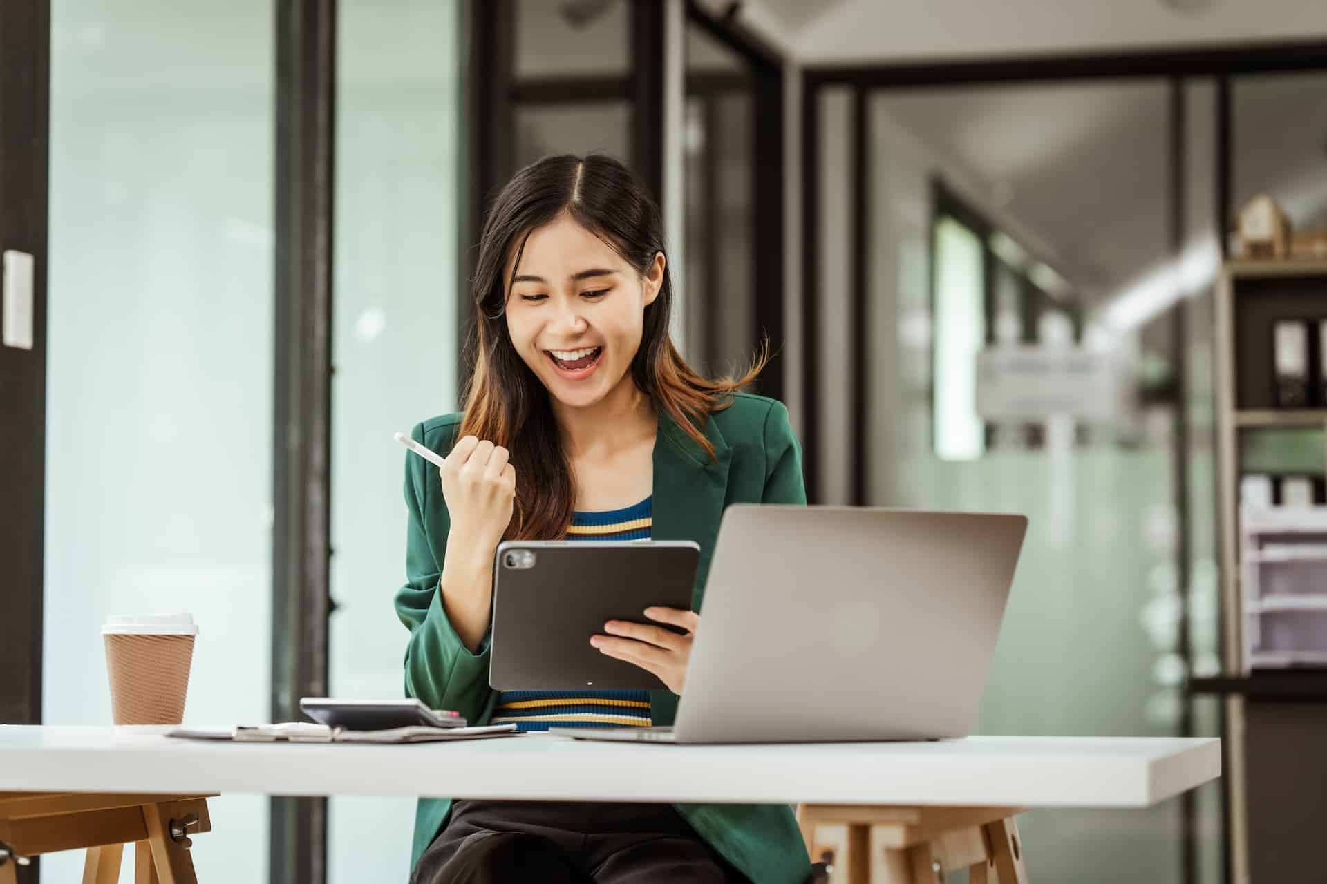 Woman in an office, looking at her iPad with a triumphant fist pump as she celebrates the success of taking a risk in her career.
