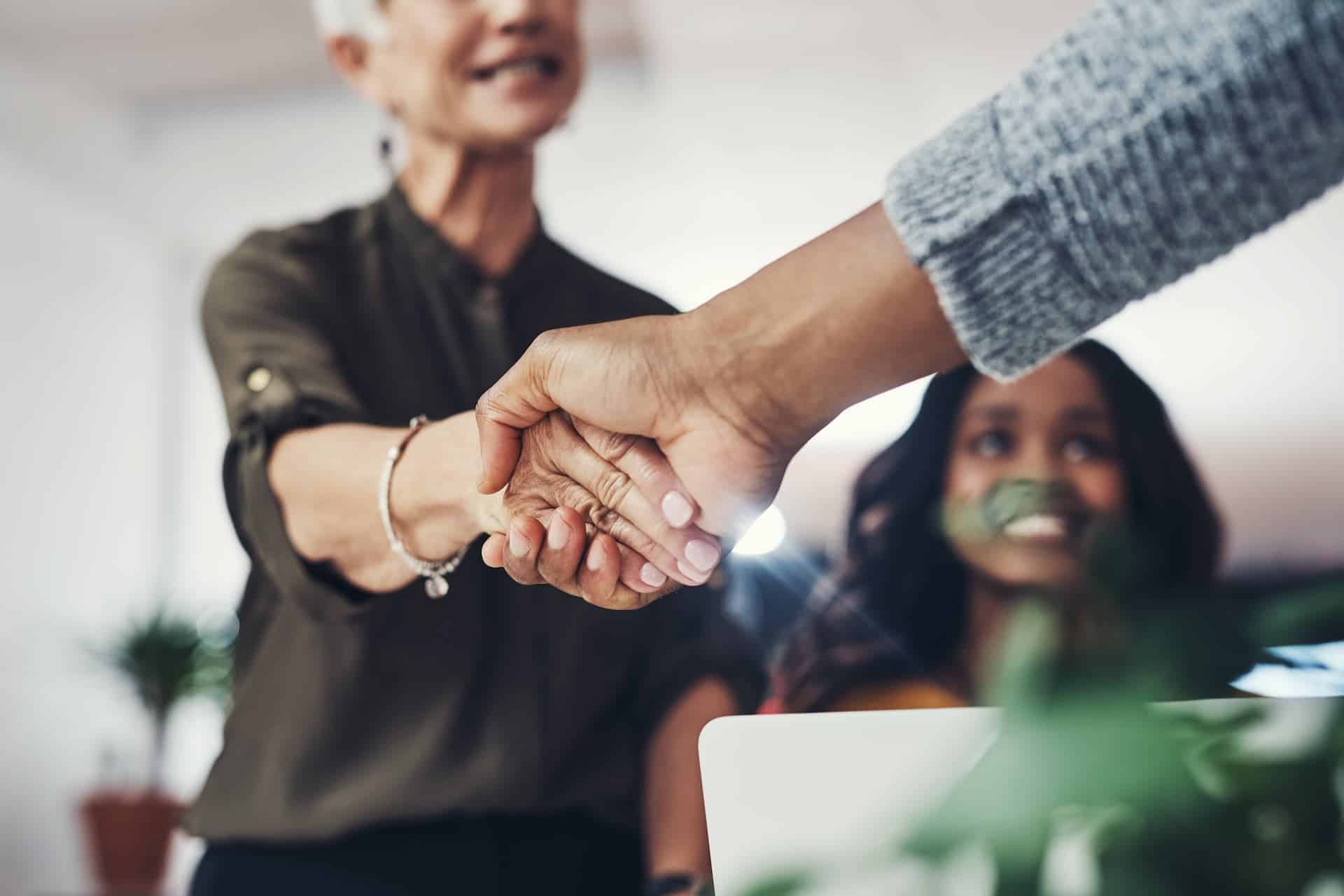 A man shaking hands with a woman, while another woman looks on with a smile, representing trust, respect, and ethical interpersonal relationships.