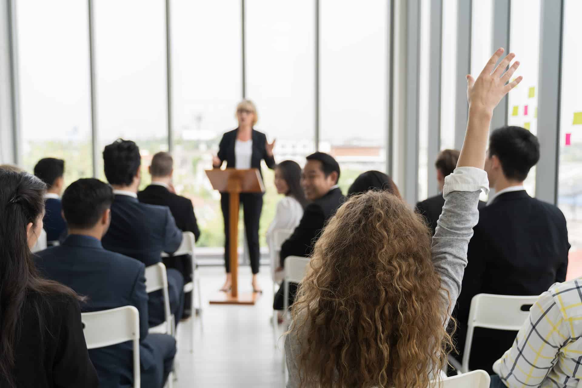 A woman raising her hand from the audience during a speech, demonstrating self-advocacy by participating actively.