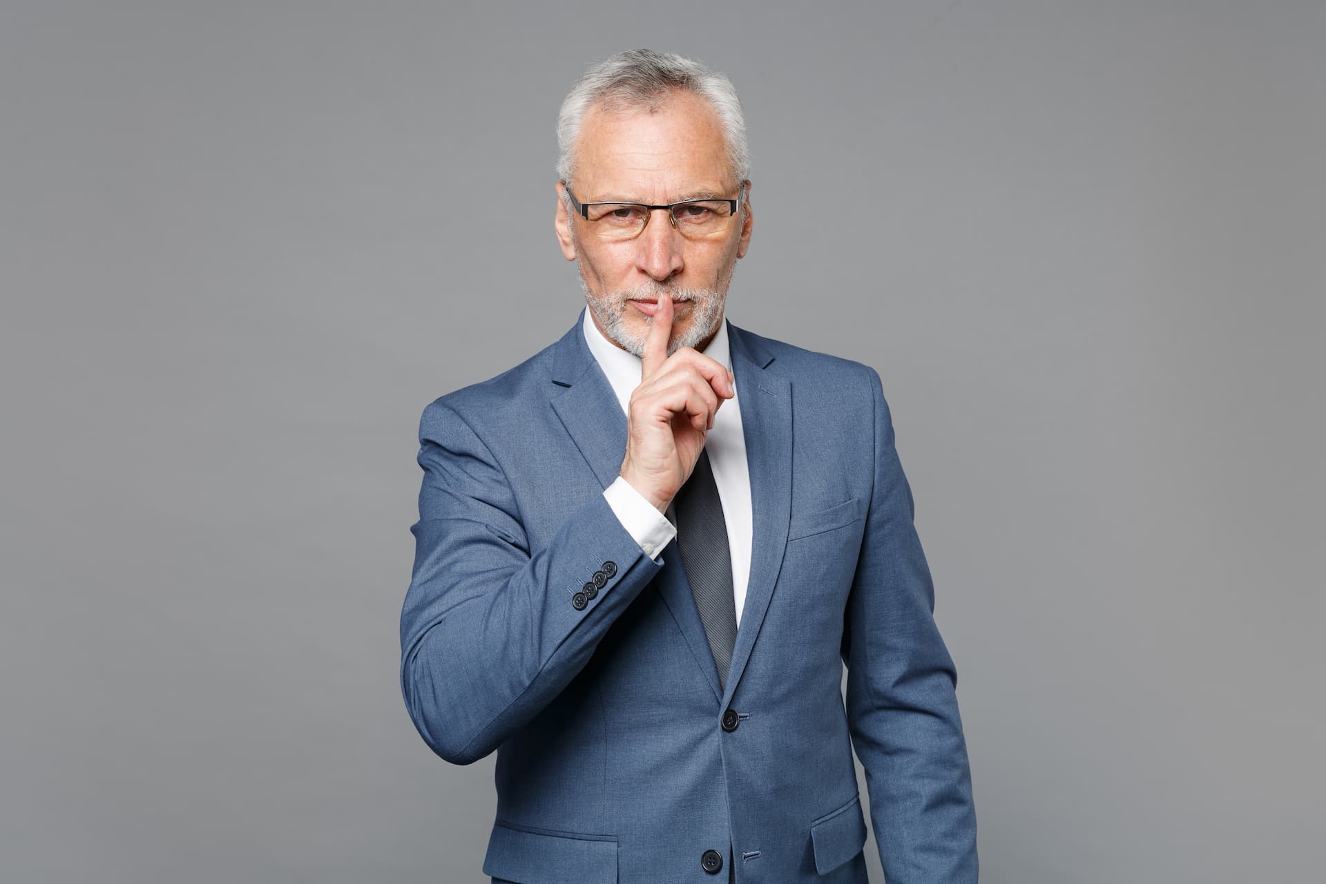 A gray-haired, bearded businessman in a blue suit, shirt, and tie, isolated against a gray background, holds his finger to his lips, signaling for silence.