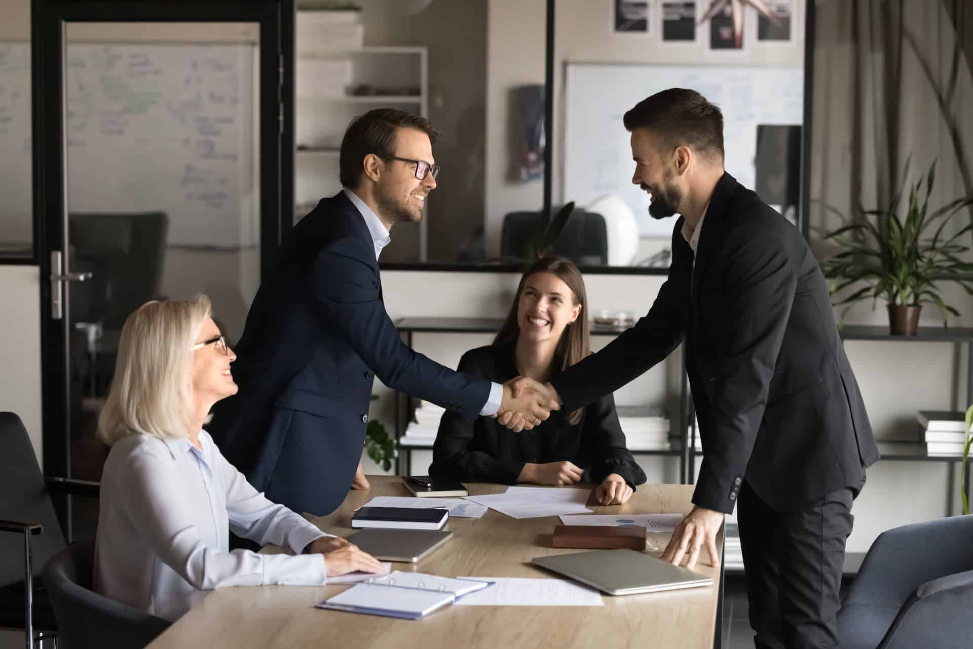 Four professionals in a conference room during a successful negotiation: two men shake hands across a table, with two women seated on either side, all smiling with satisfaction.
