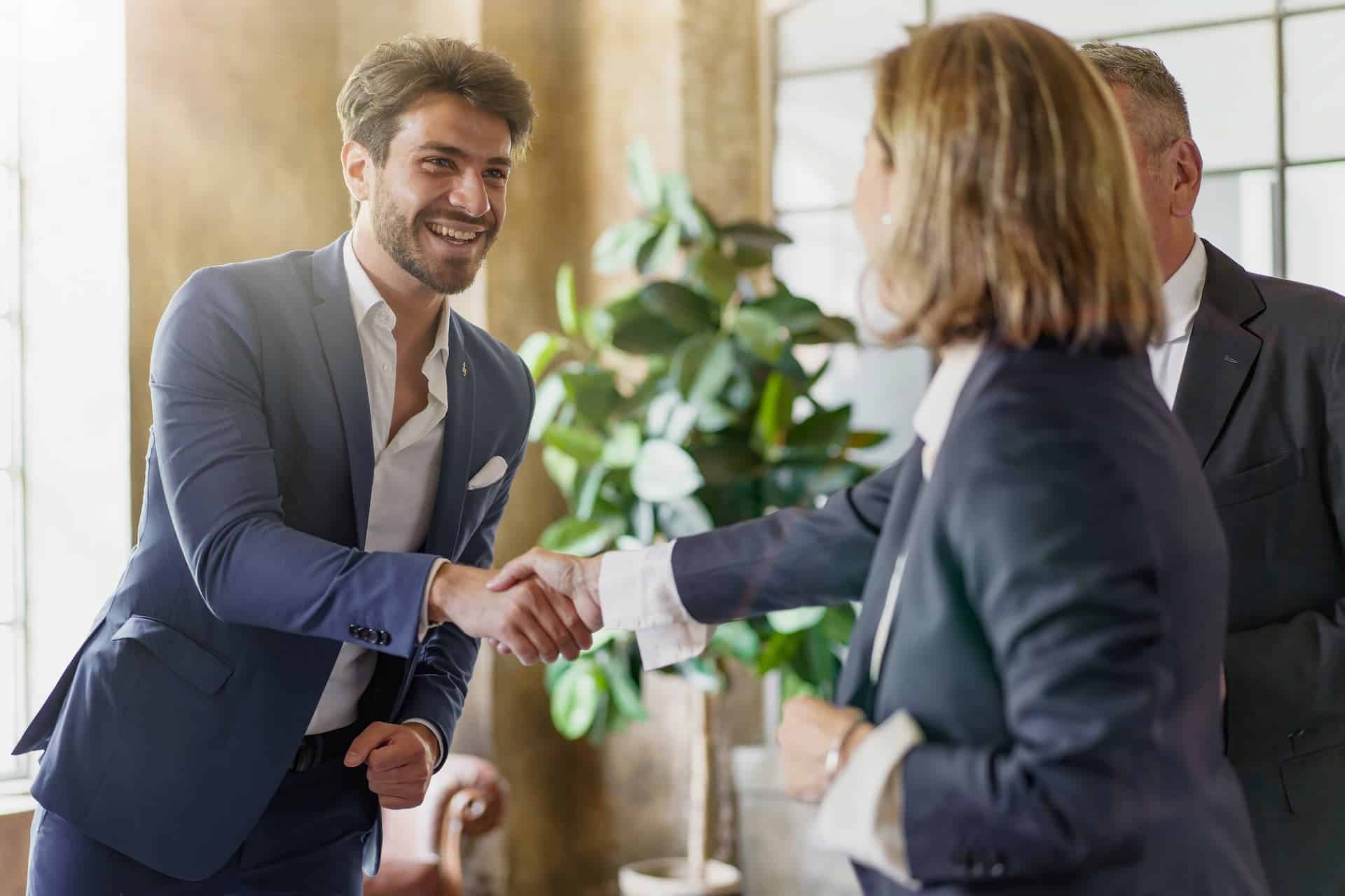 A man smiling while networking, shaking hands with a woman and another man, confidently relying on body language that conveys friendliness and professionalism.
