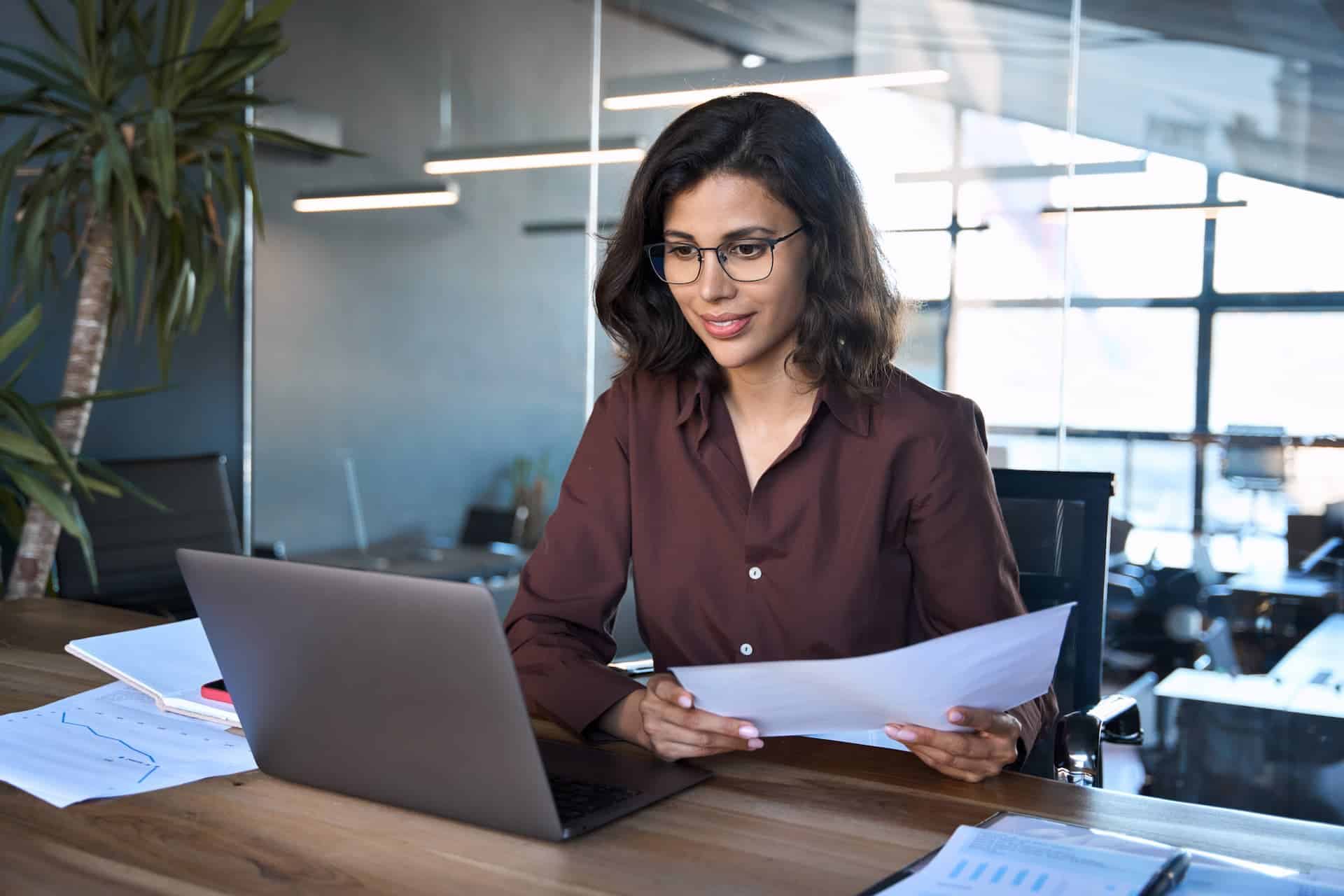 A woman sitting at a desk, holding a paper while reviewing her work on her laptop, embodying the reflective process of deliberate practice to enhance her skills.