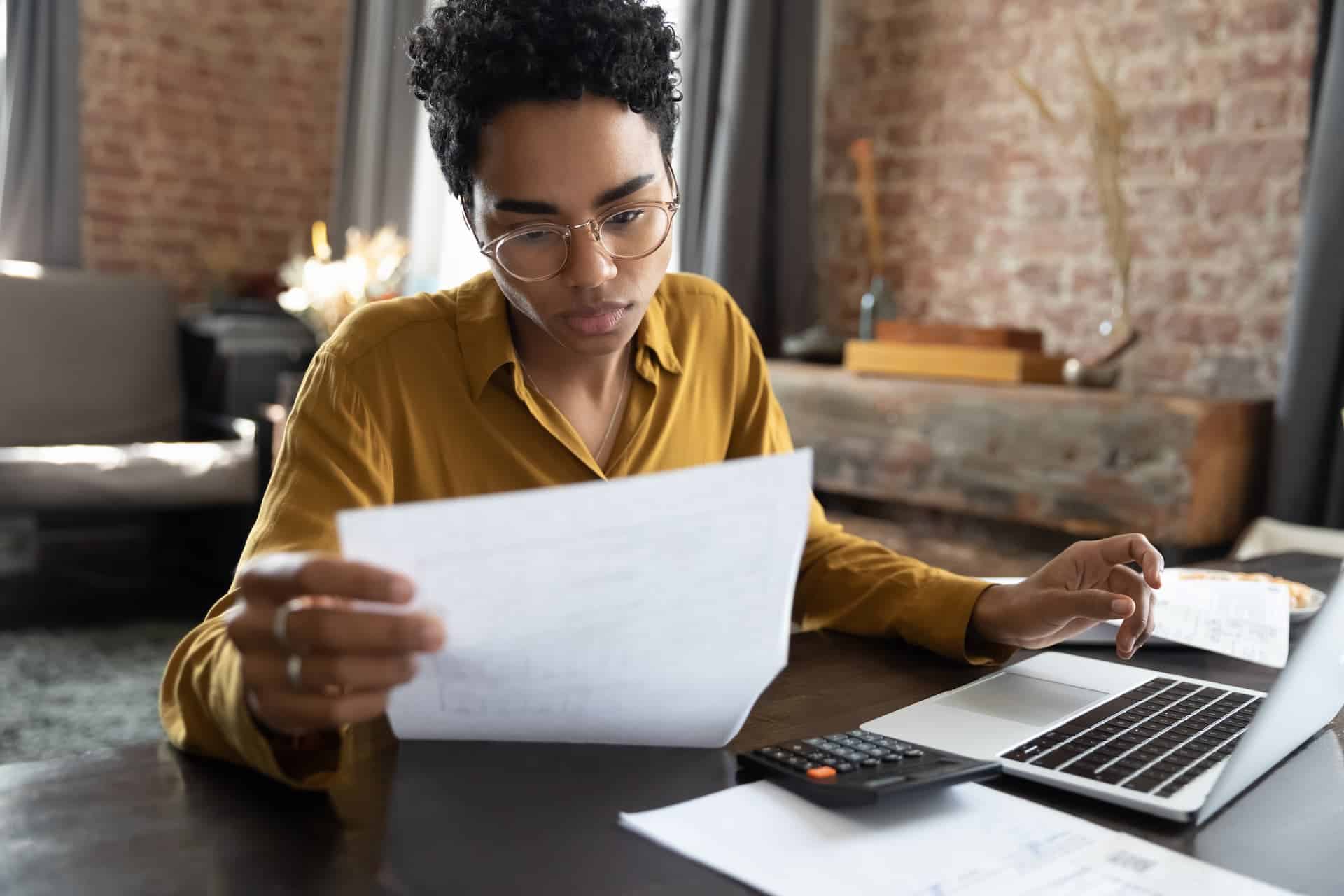 A woman engaged in deliberate practice, holding a sheet of paper and focusing on her laptop, illustrating a study session aimed at skill improvement.