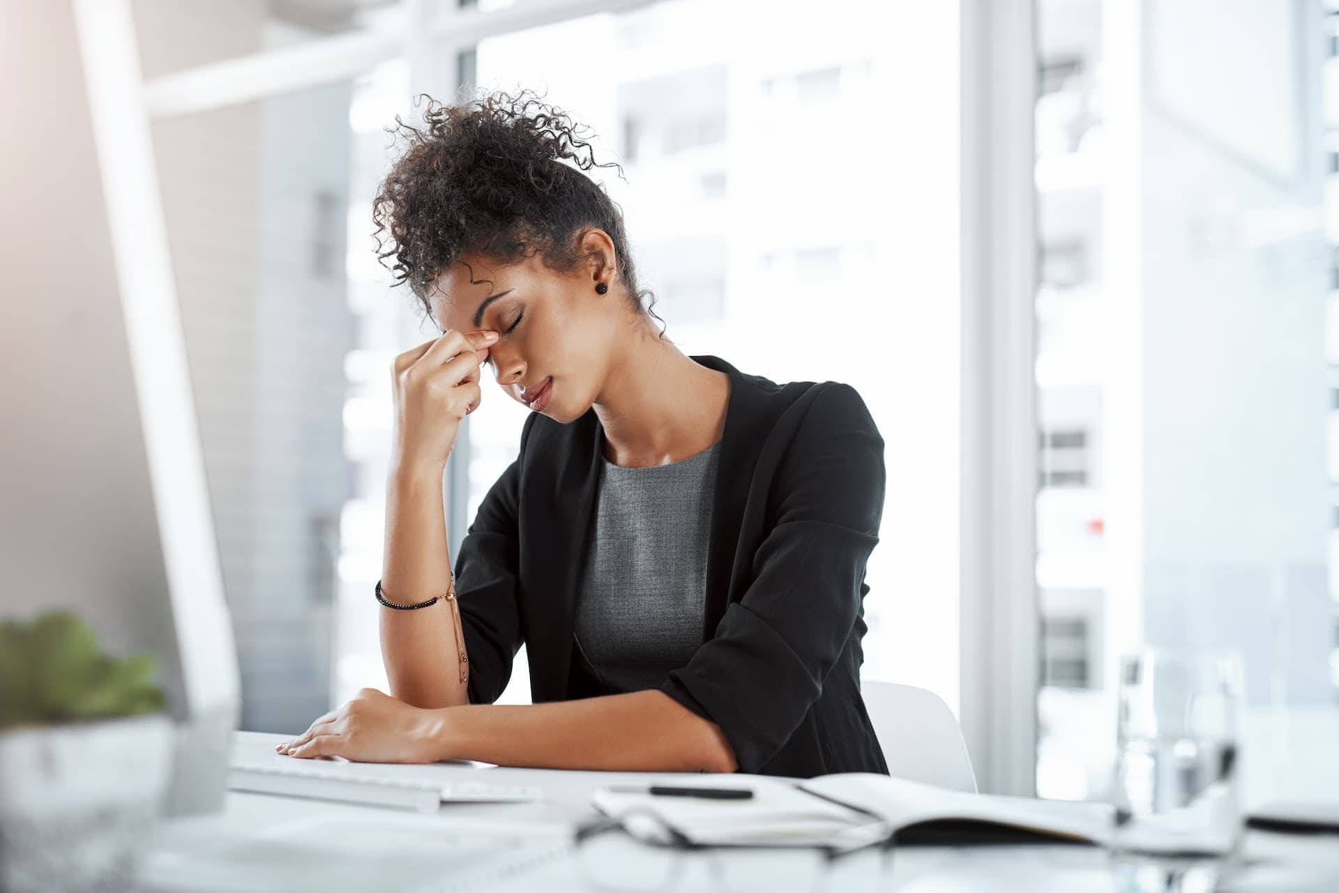 A businesswoman, clearly experiencing burnout, sits at her desk in an office, eyes closed as she pinches the bridge of her nose.