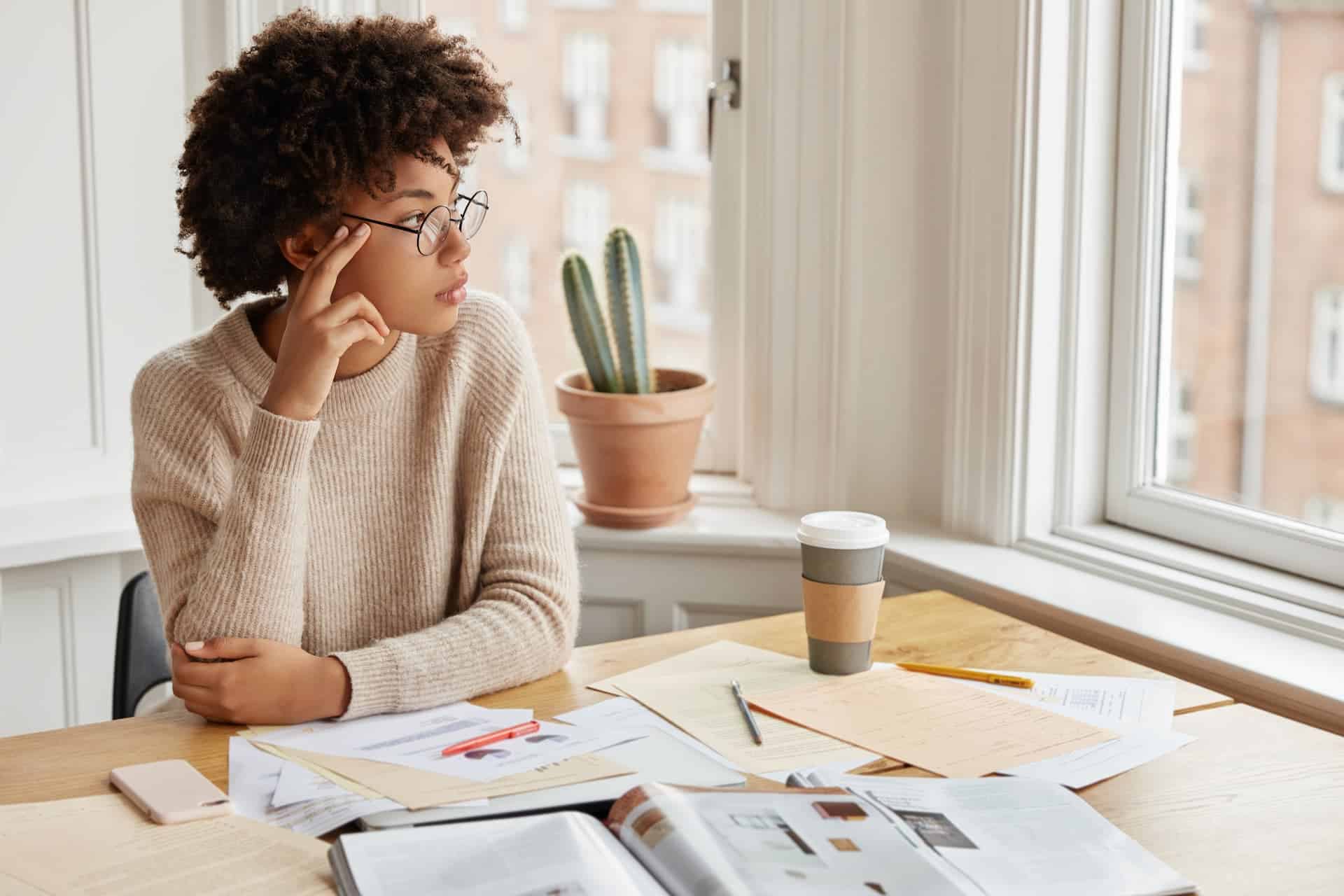A woman looking out the window while books are open on her table, showing a thoughtful moment of reflection and highlighting the tenacity needed to gain knowledge and understanding.