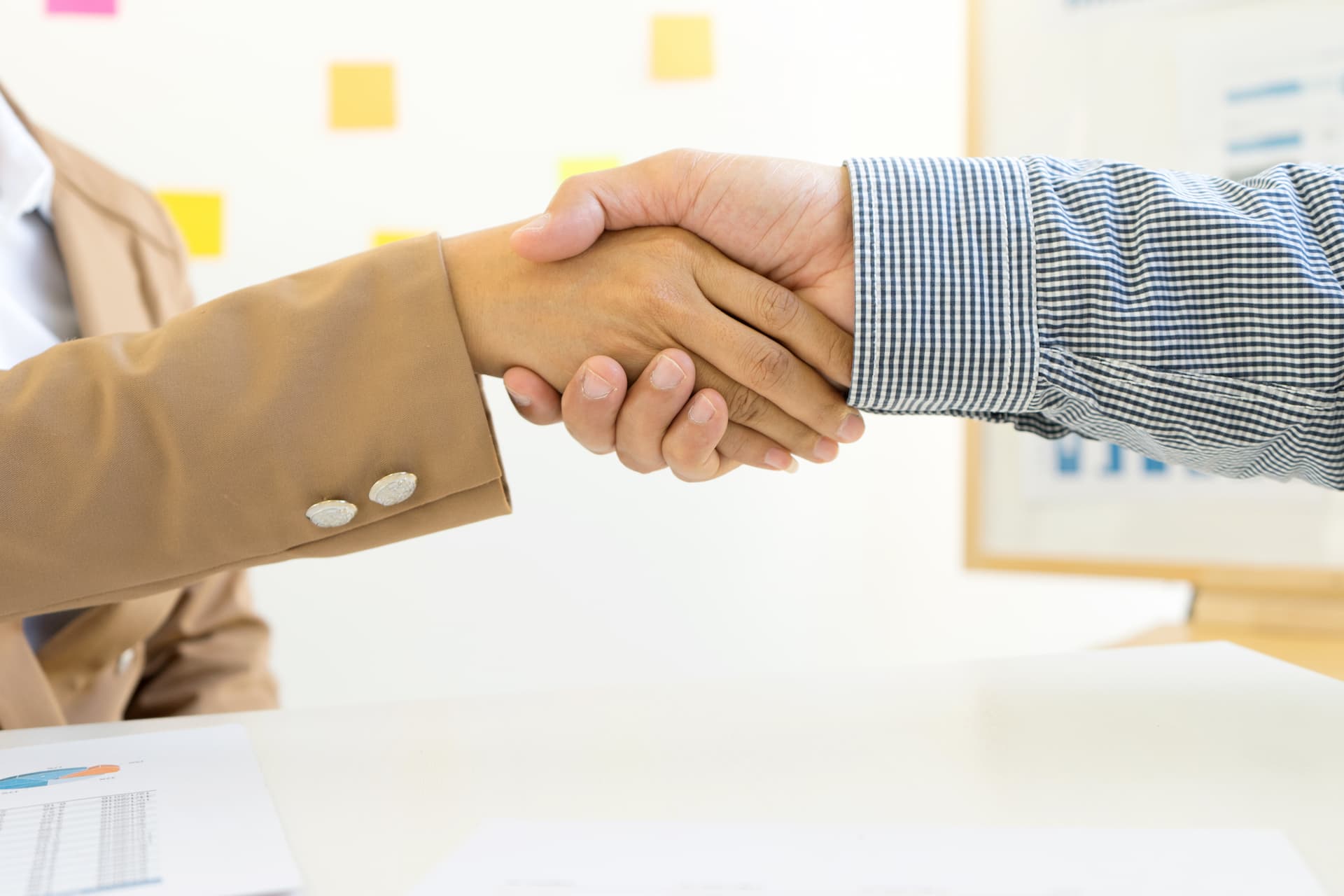 Close-up of two hands shaking, with forearms visible just beyond the wrists, set against a blurred office background.