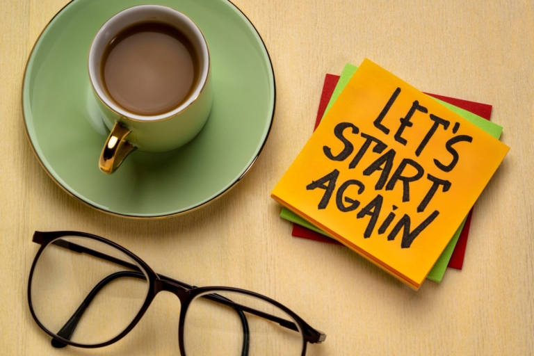 A coffee mug with coffee, glasses, and a post-it note that reads 'Let's start again' - a setup symbolizing the determination to start anew and persist through challenges.
