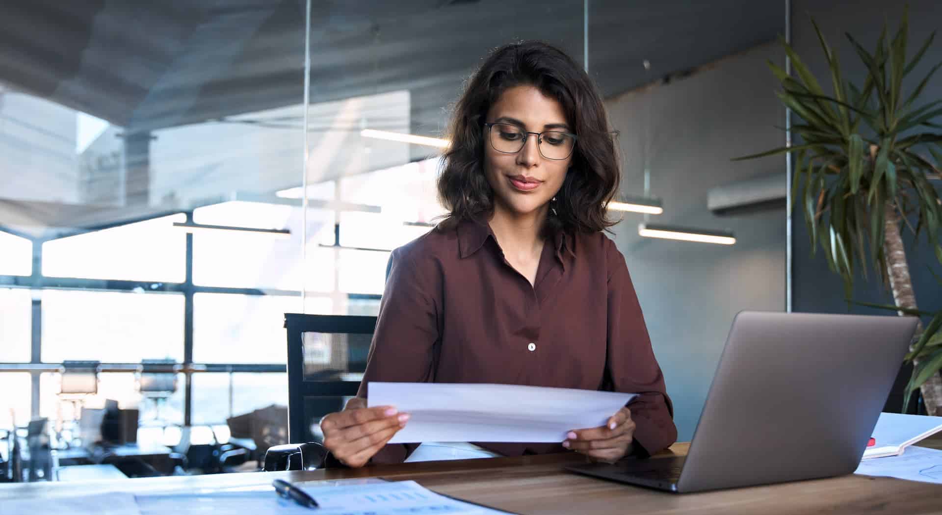 A woman in an office, focused on a document with her laptop open in front of her, making sure her work is consistent. 