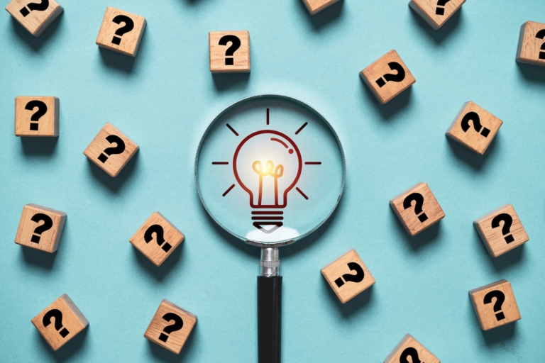 Several dice with question marks on them and a magnifying glass at the center showing a lightbulb, illustrating the concept of having great ideas by asking the right questions.