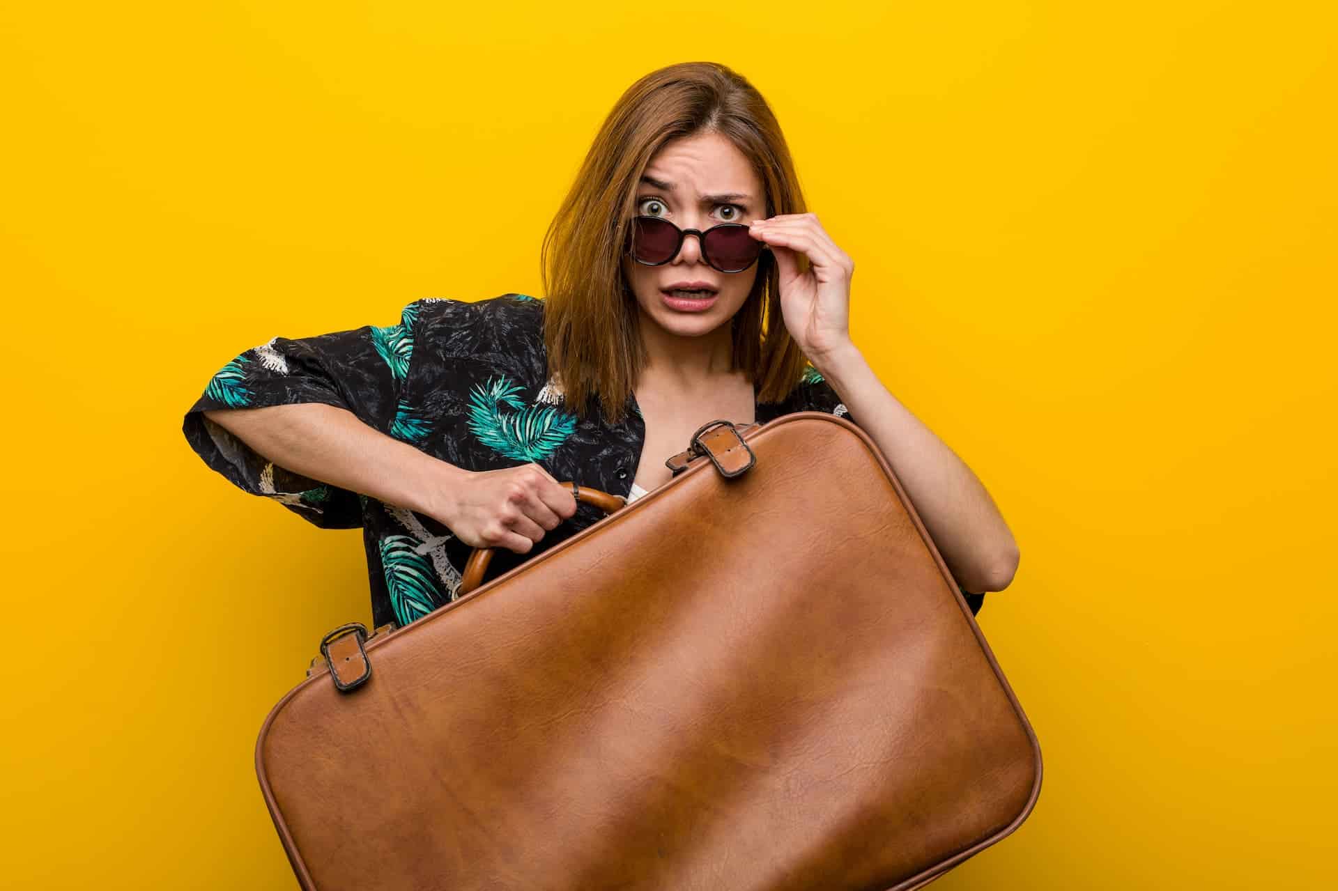 A woman in a tropical shirt looks stressed while holding a large brown suitcase in front of her, adjusting her sunglasses and contemplating her emotional baggage against a bright yellow background.