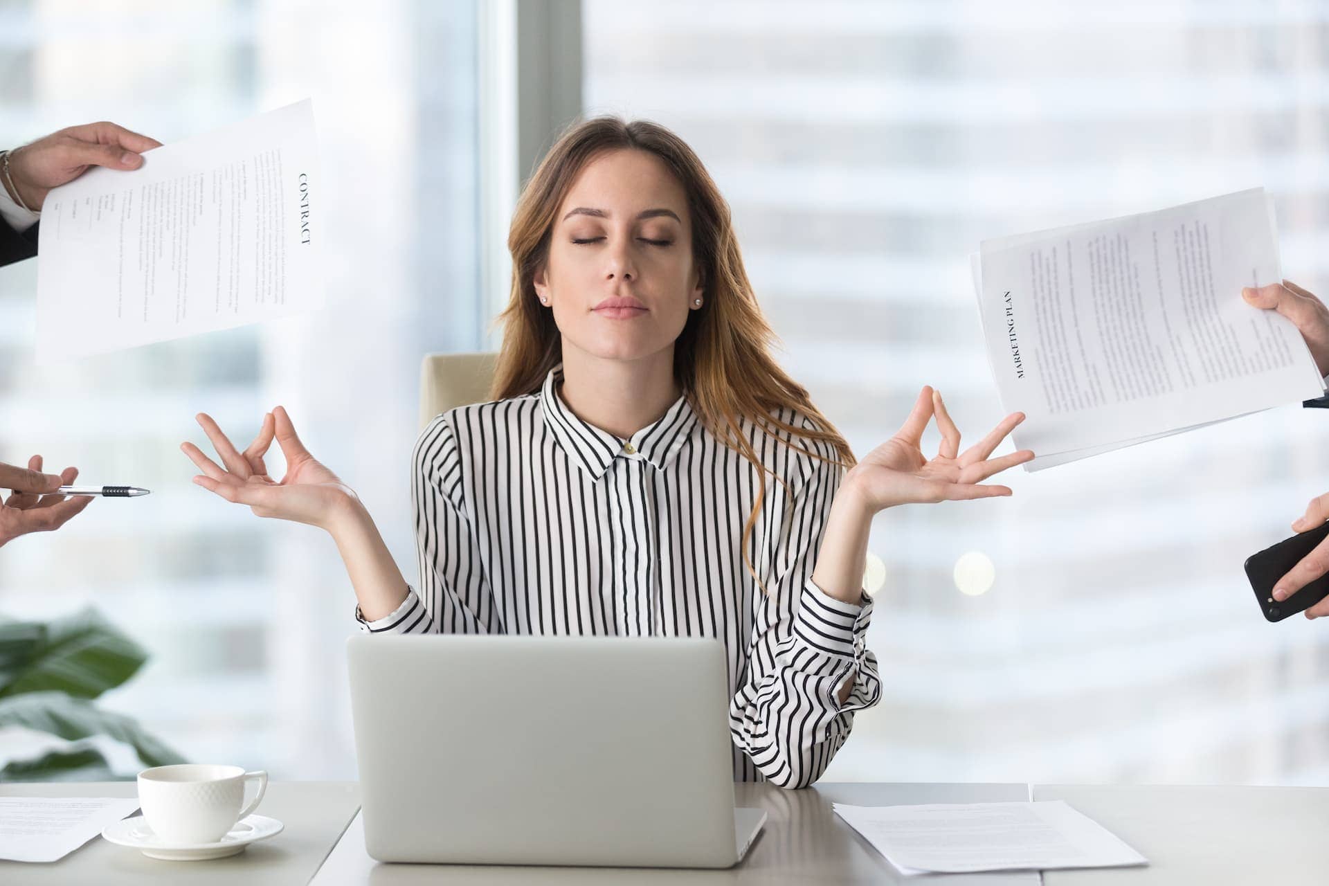 A woman meditating at her desk amidst work chaos, demonstrating stress management techniques in a busy office.