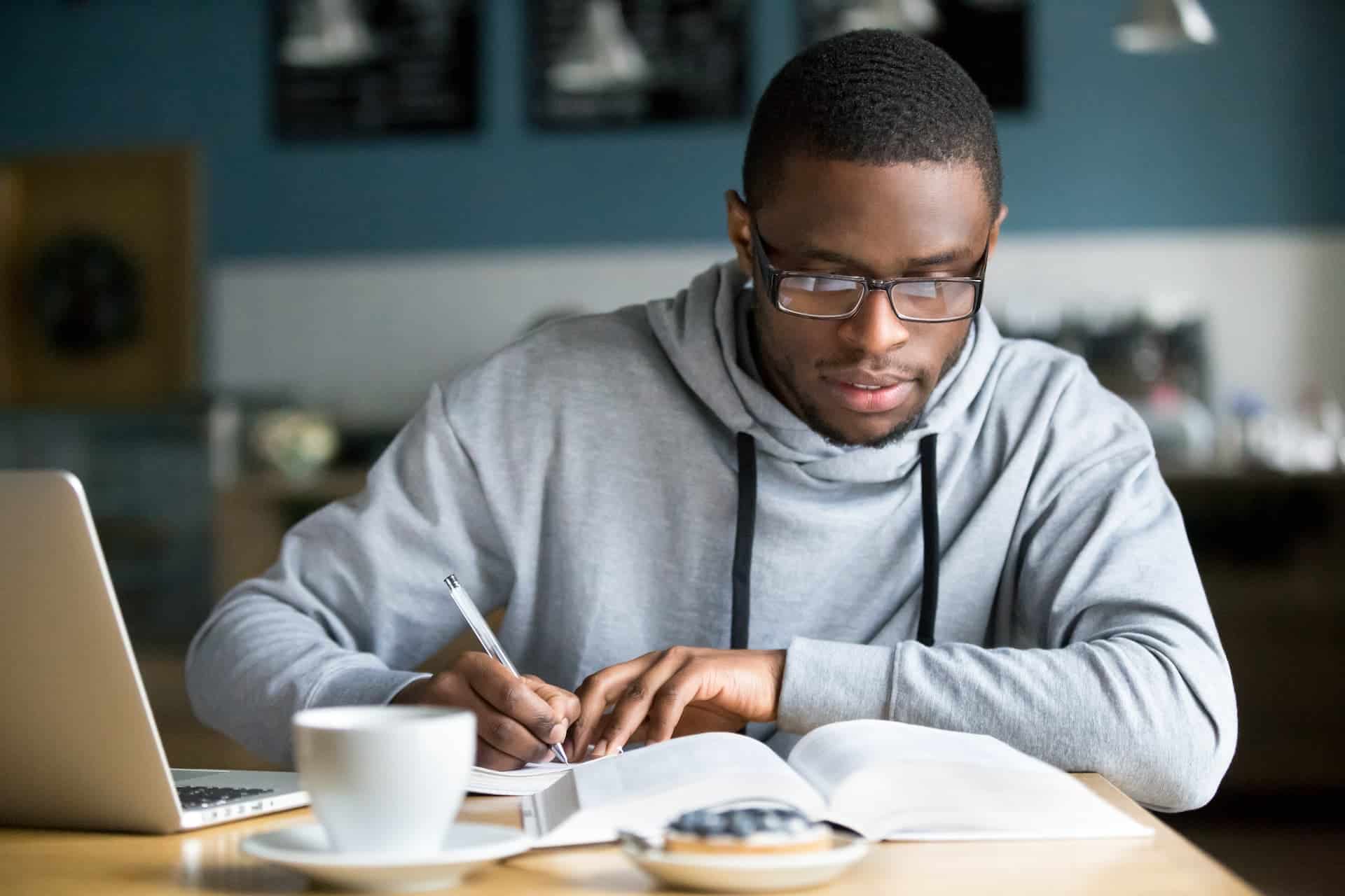 A man synthesizing information from a book while summarizing on his laptop.