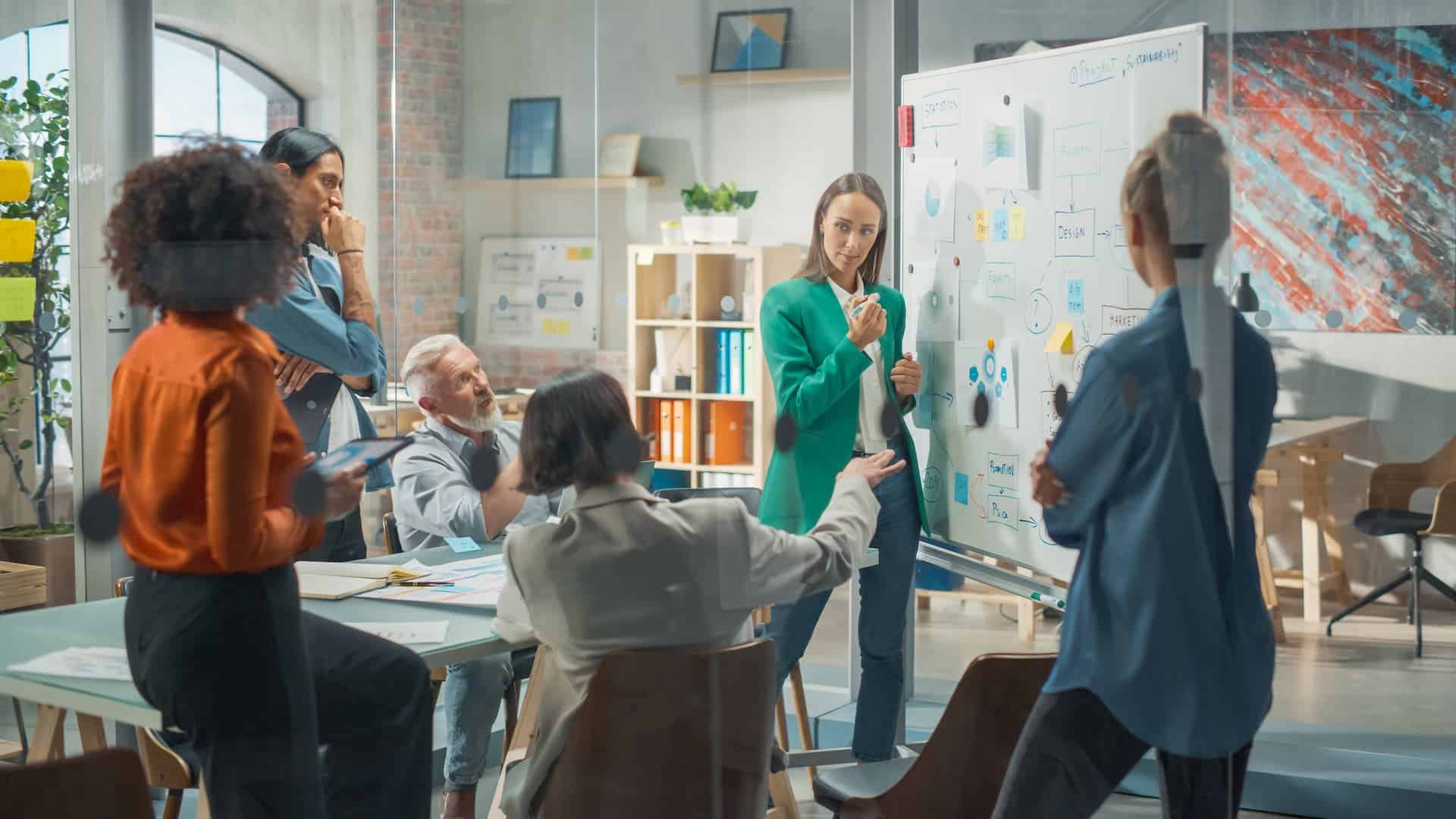 A team standing and sitting around a conference table focused on a large whiteboard while having a brainstorming session.